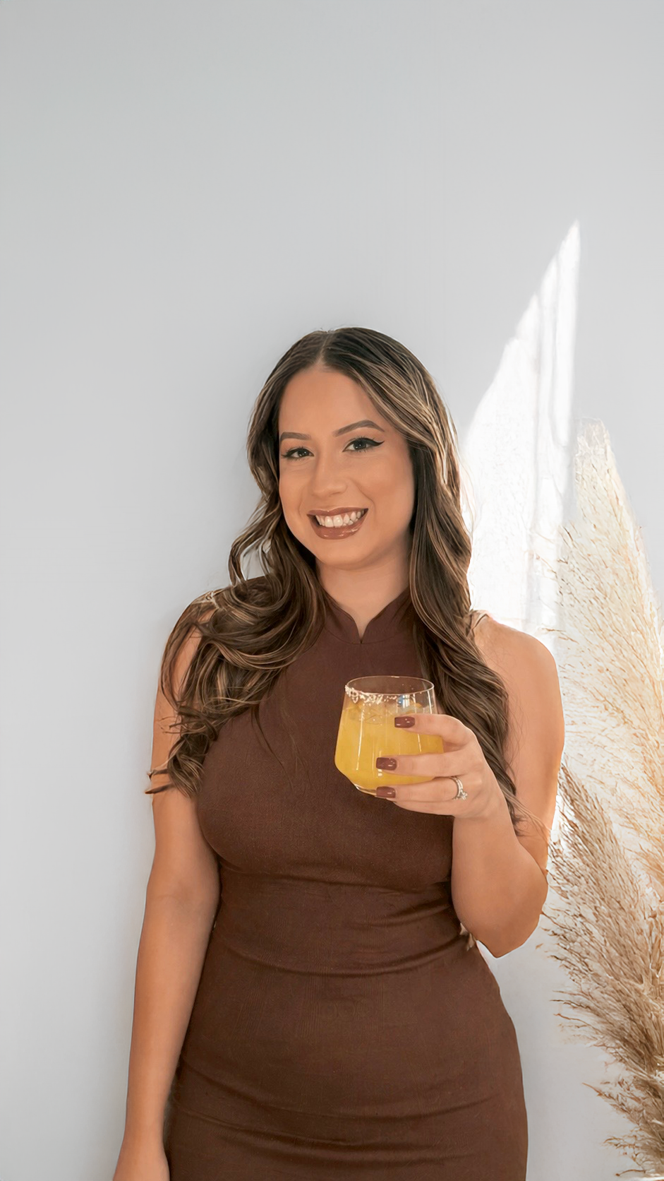 A woman in a brown sleeveless dress holding a cocktail beverage, smiling, with a beige decorative plant behind her.