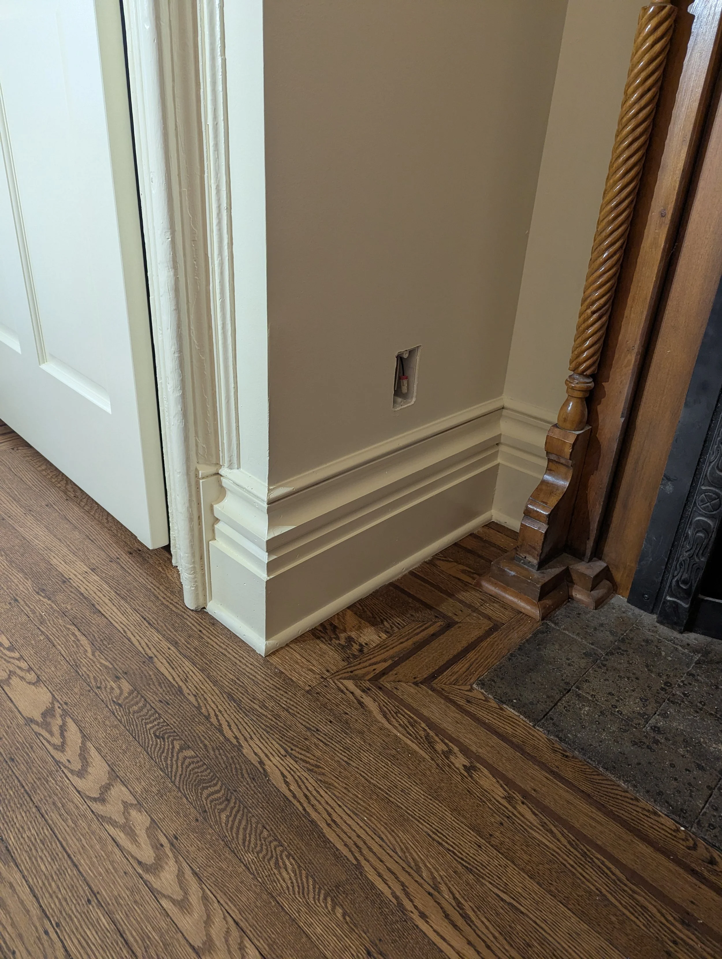 Corner of a room with hardwood flooring, a painted wall with a small electrical outlet, and a wooden staircase railing.
