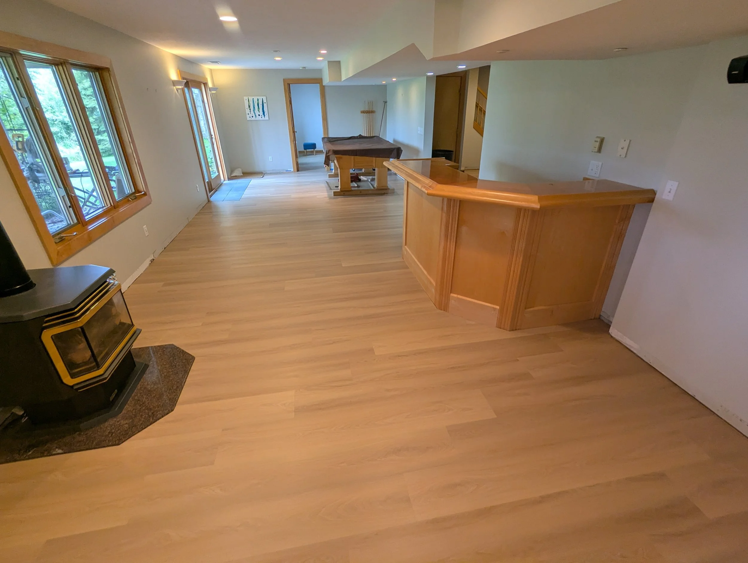 Living room with hardwood floors, large windows with wooden frames, a small grand piano on a rug, a doorway, art on the wall, a pool cue rack, and a pool table covered with a brown cloth.