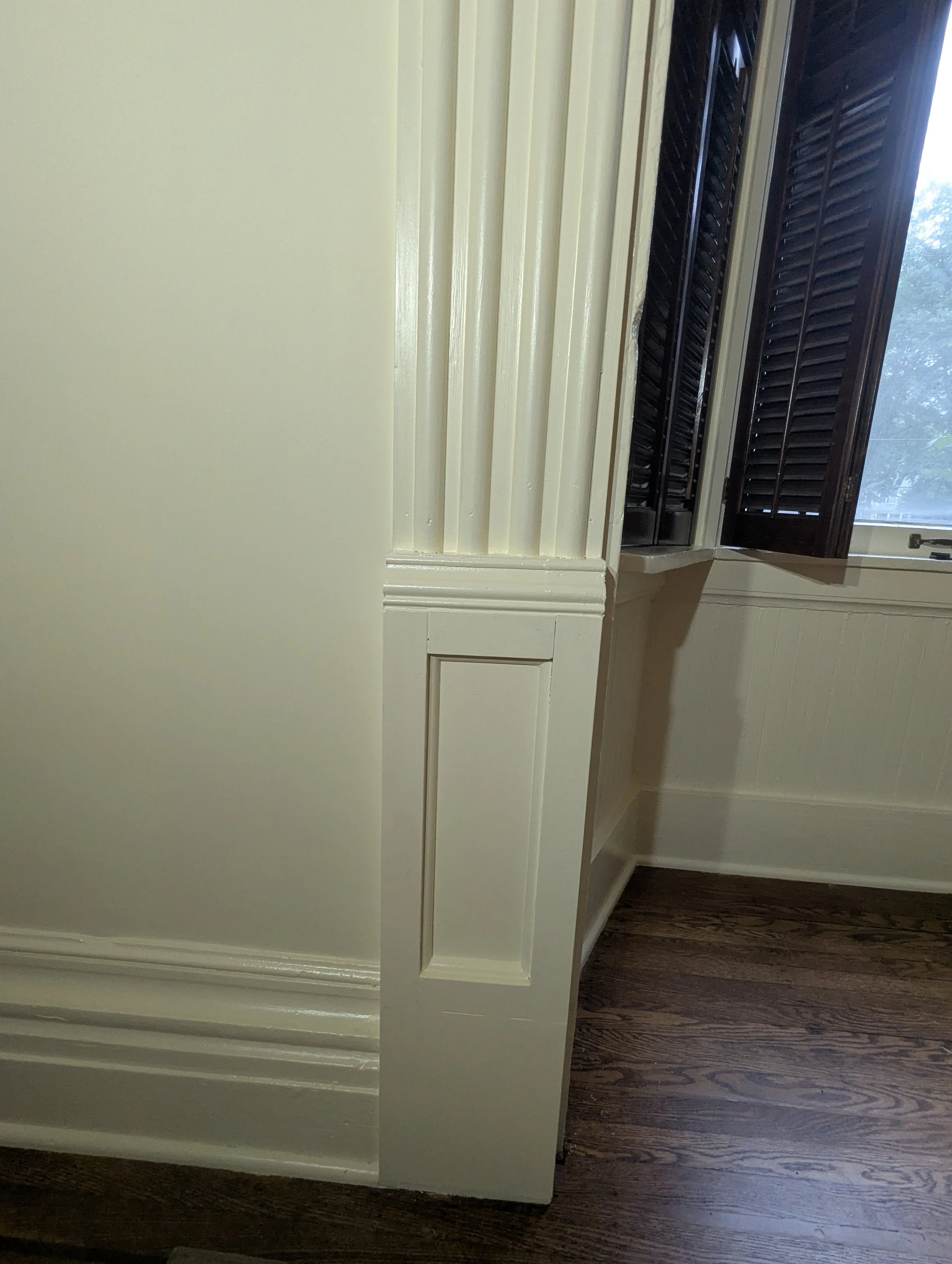 Interior view of a room corner with a window with dark shutters, beige walls, ornate white trim, and dark wooden flooring.