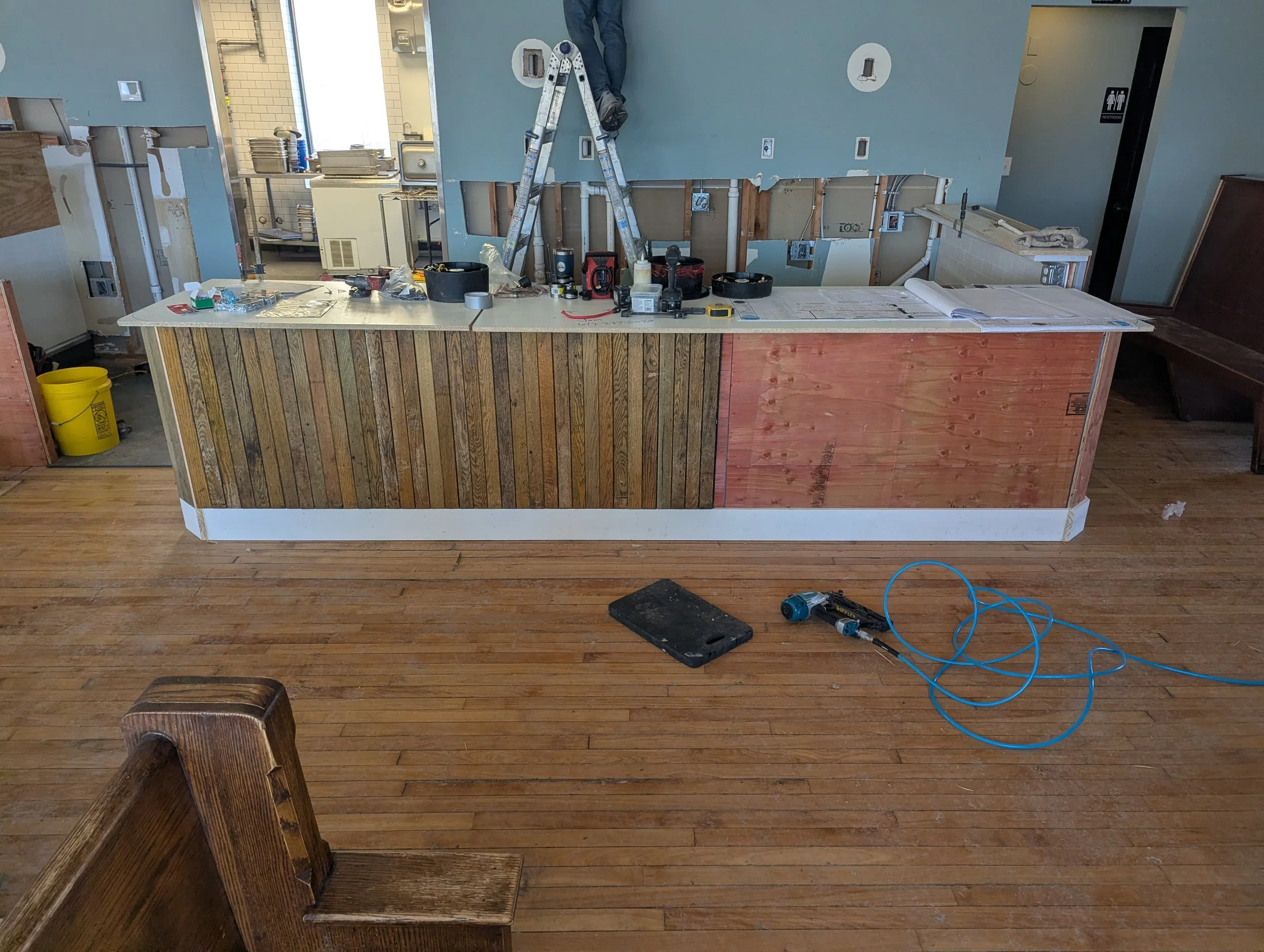 Interior of a restaurant under renovation with a counter made of wood panels, tools, and construction materials scattered on top, an open wall revealing wiring and plumbing, a ladder with a worker, and a man in the background working near a window.