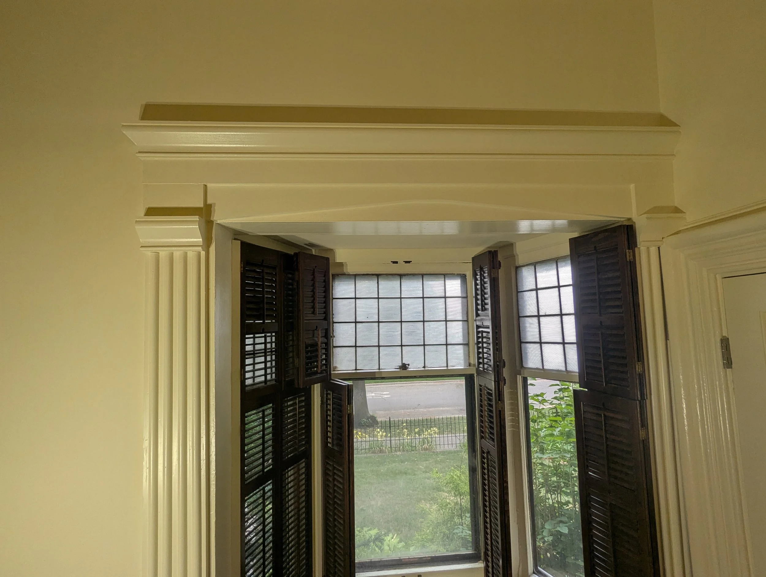View of an interior room with tall windows and dark wooden shutters, looking outside to a garden with grass and plants.