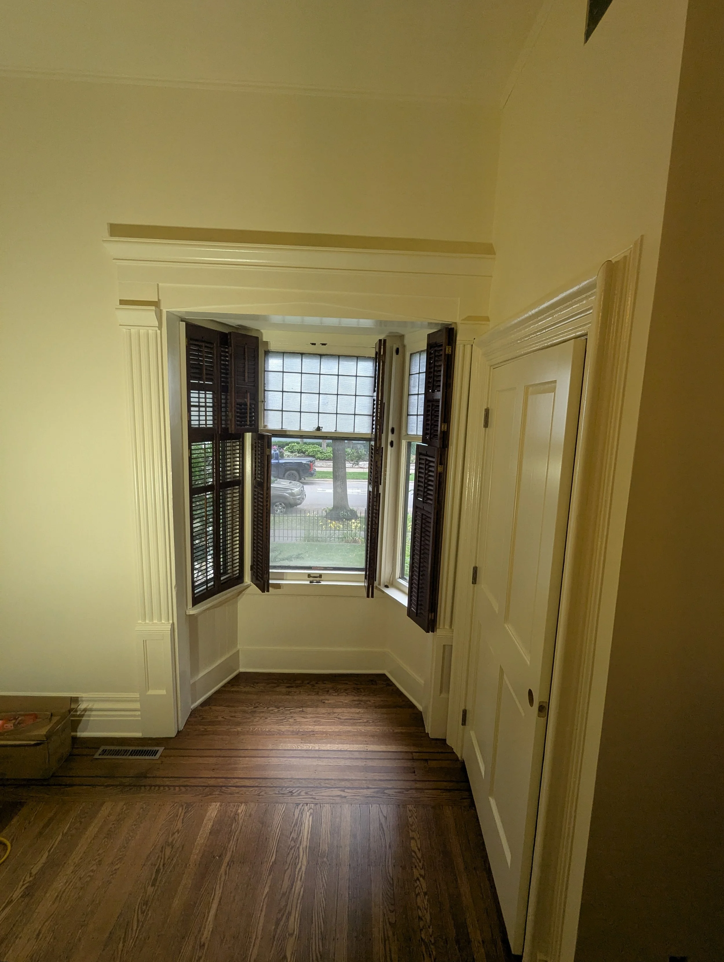 A room with a large window, white walls, dark wooden shutters, and hardwood flooring.