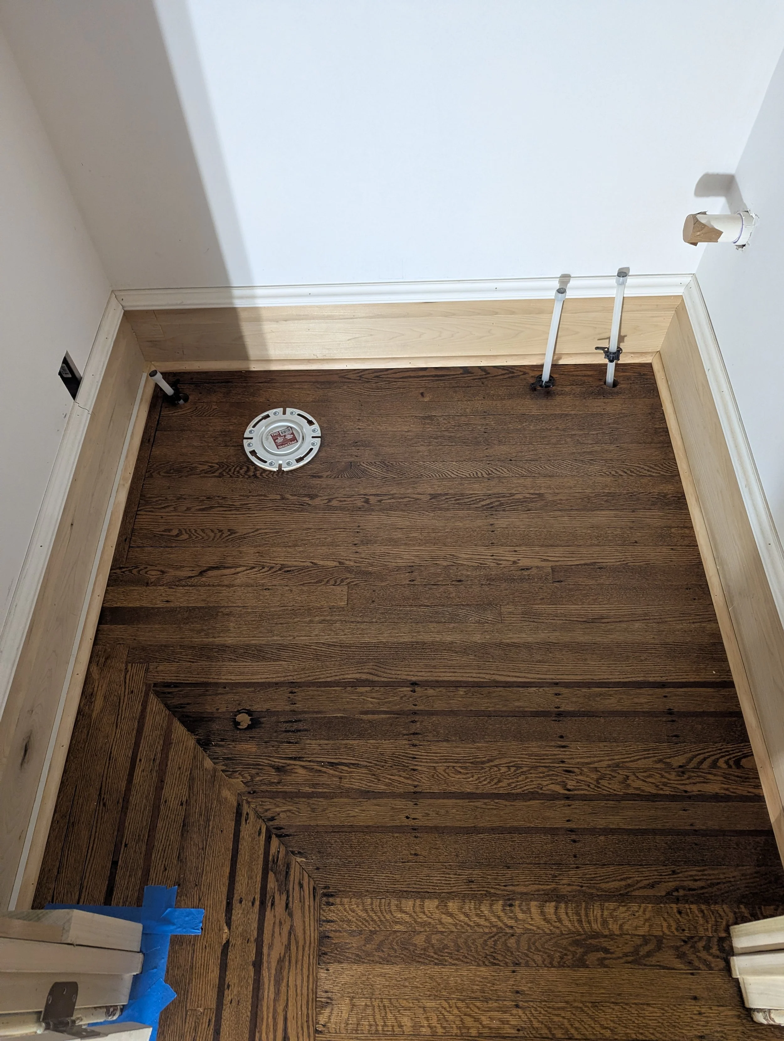 A corner of a room with hardwood flooring, white walls, and some wall repairs in progress. There are pipes and a smoke detector on the floor, and blue painter's tape on the lower left corner.