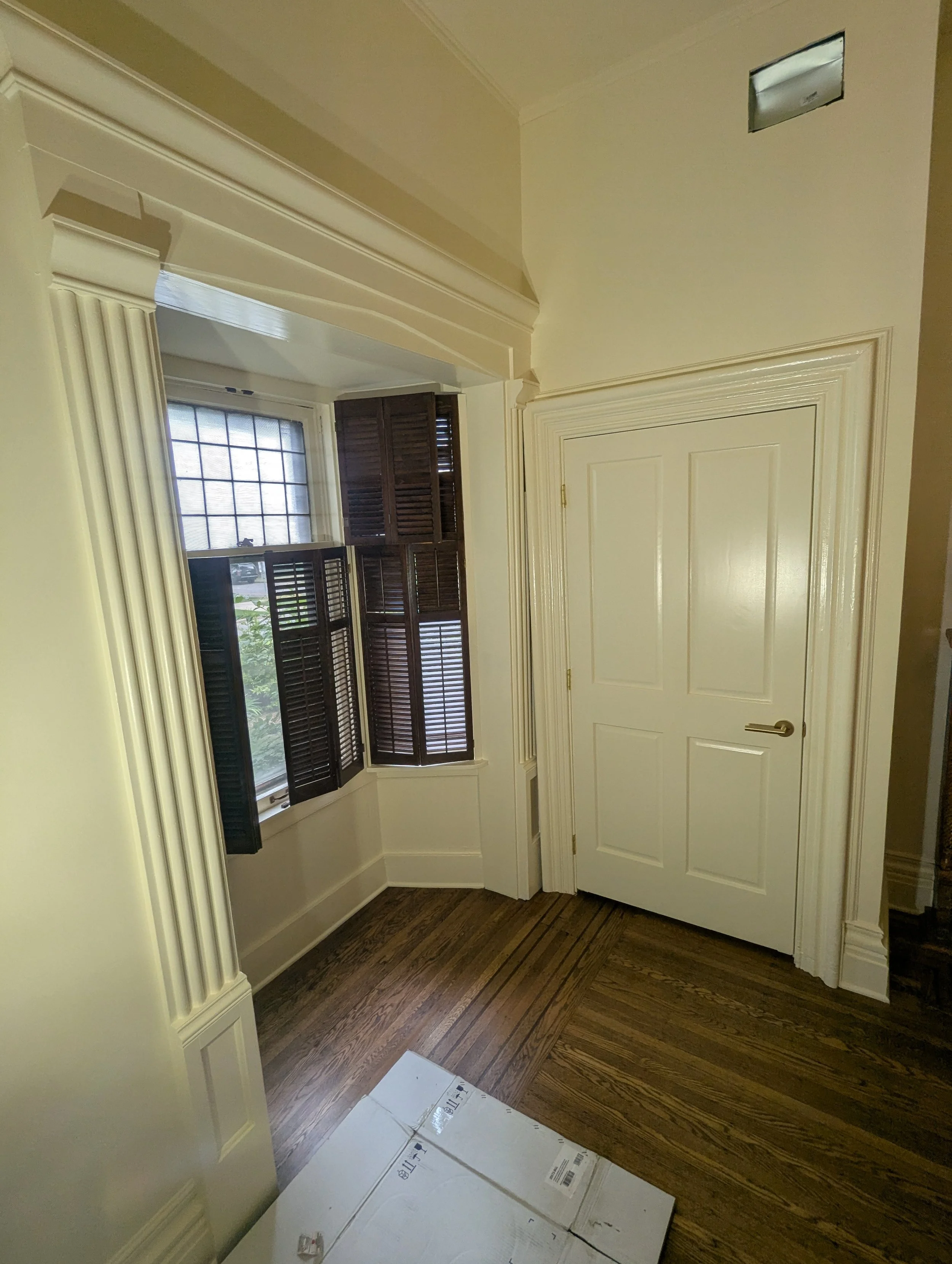 A room with cream-colored walls and an open door. There are dark wooden blinds on the window and a cardboard box on the floor near the door.