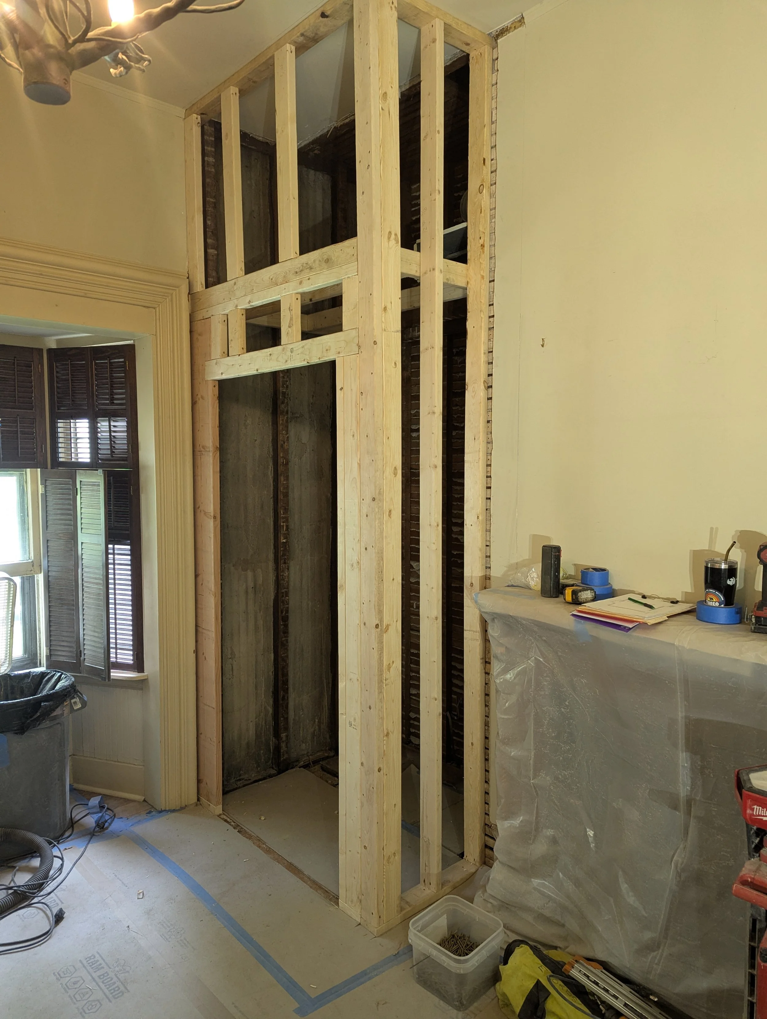 Interior of a room under renovation with a wooden framing structure, a window with shutters, tools, and construction materials visible.