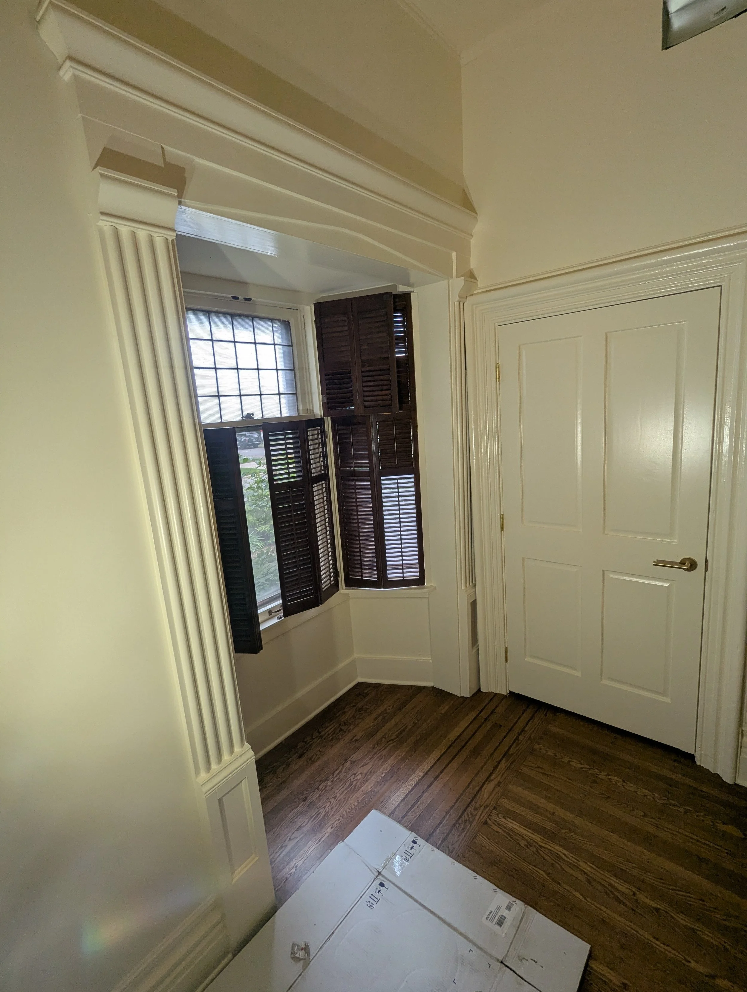 An interior view of a room showing a window with dark shutters, a closed door with a gold handle, wooden flooring, and some boxes on the floor near the door.