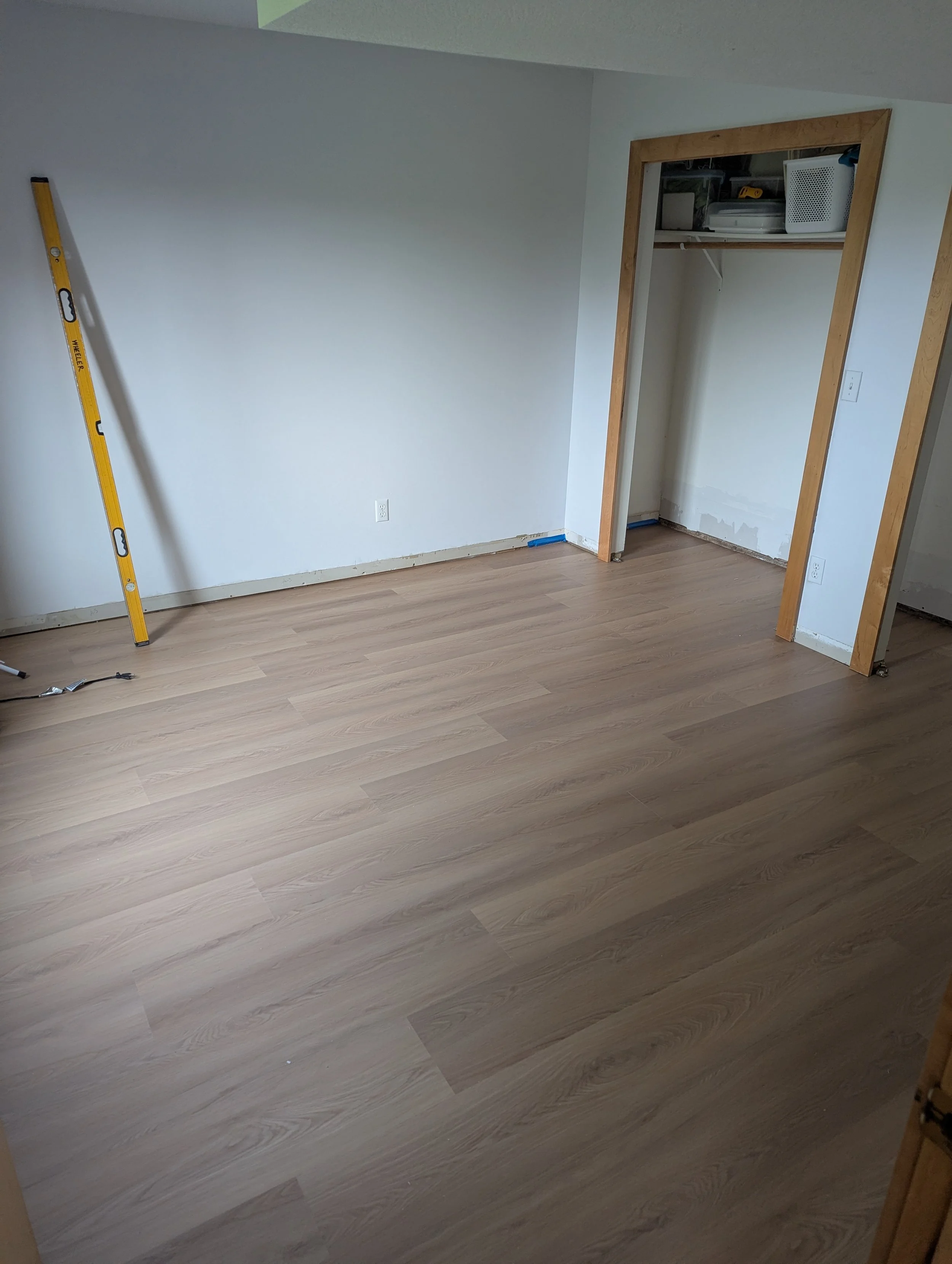 Room with new light wood flooring, an open closet with some storage boxes and items on a shelf, a yellow level leaning against the wall, and some tools on the floor.