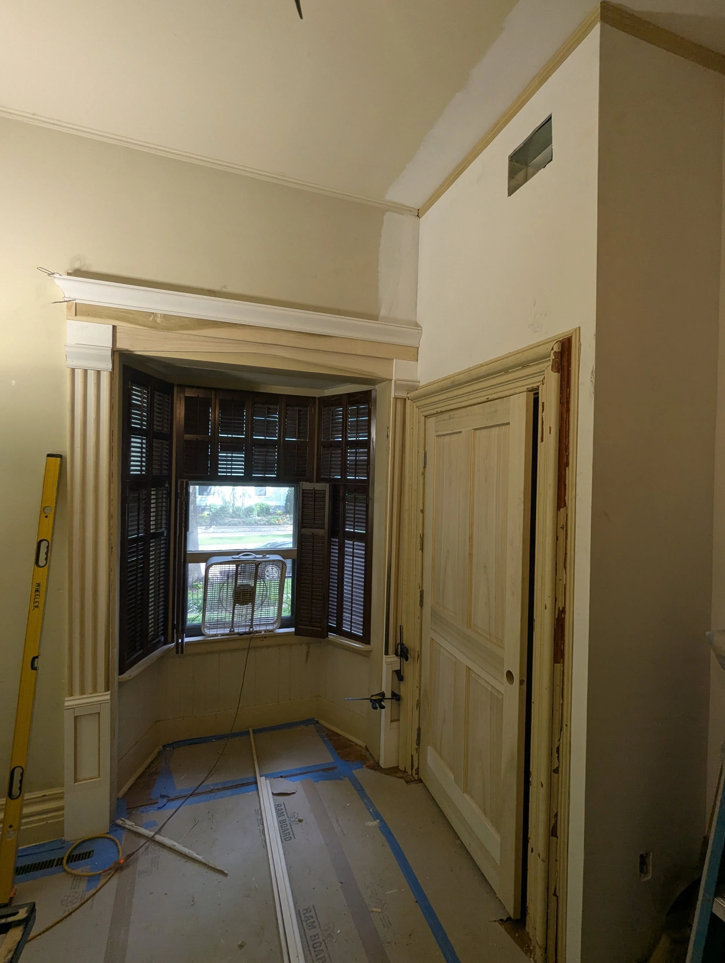 Interior of a room under construction or renovation, with partially installed wooden cabinets, a window with shutters, and construction tools and materials on the floor.