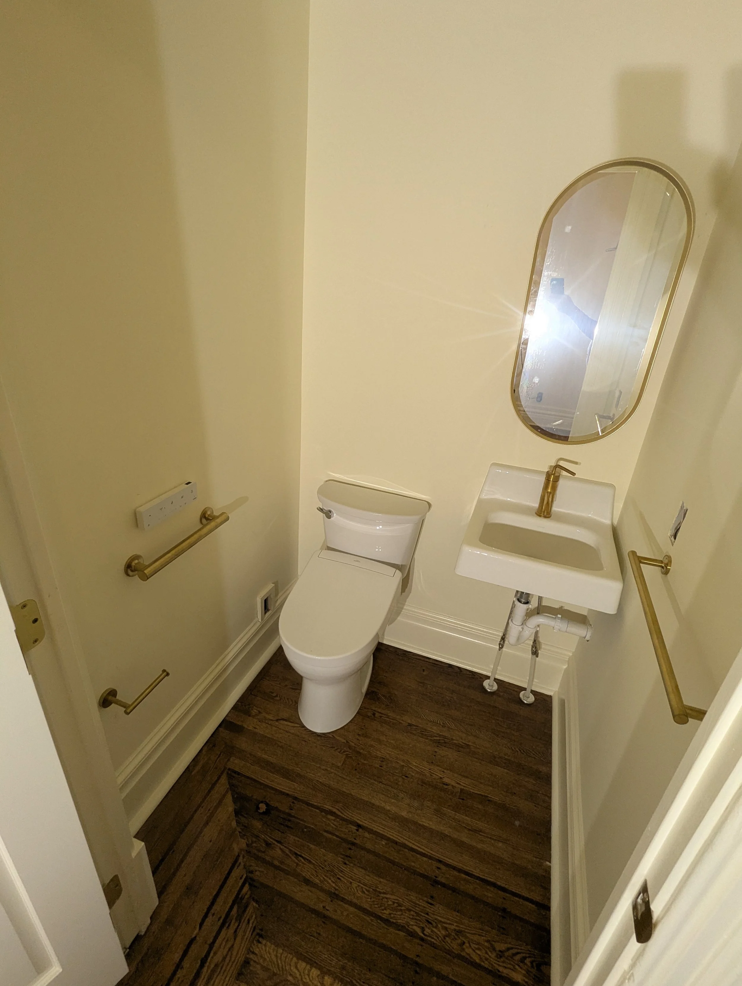 Small bathroom with yellow walls, wooden floor, a toilet, a small sink with a gold faucet, an oval mirror with a gold frame, and gold grab bars.