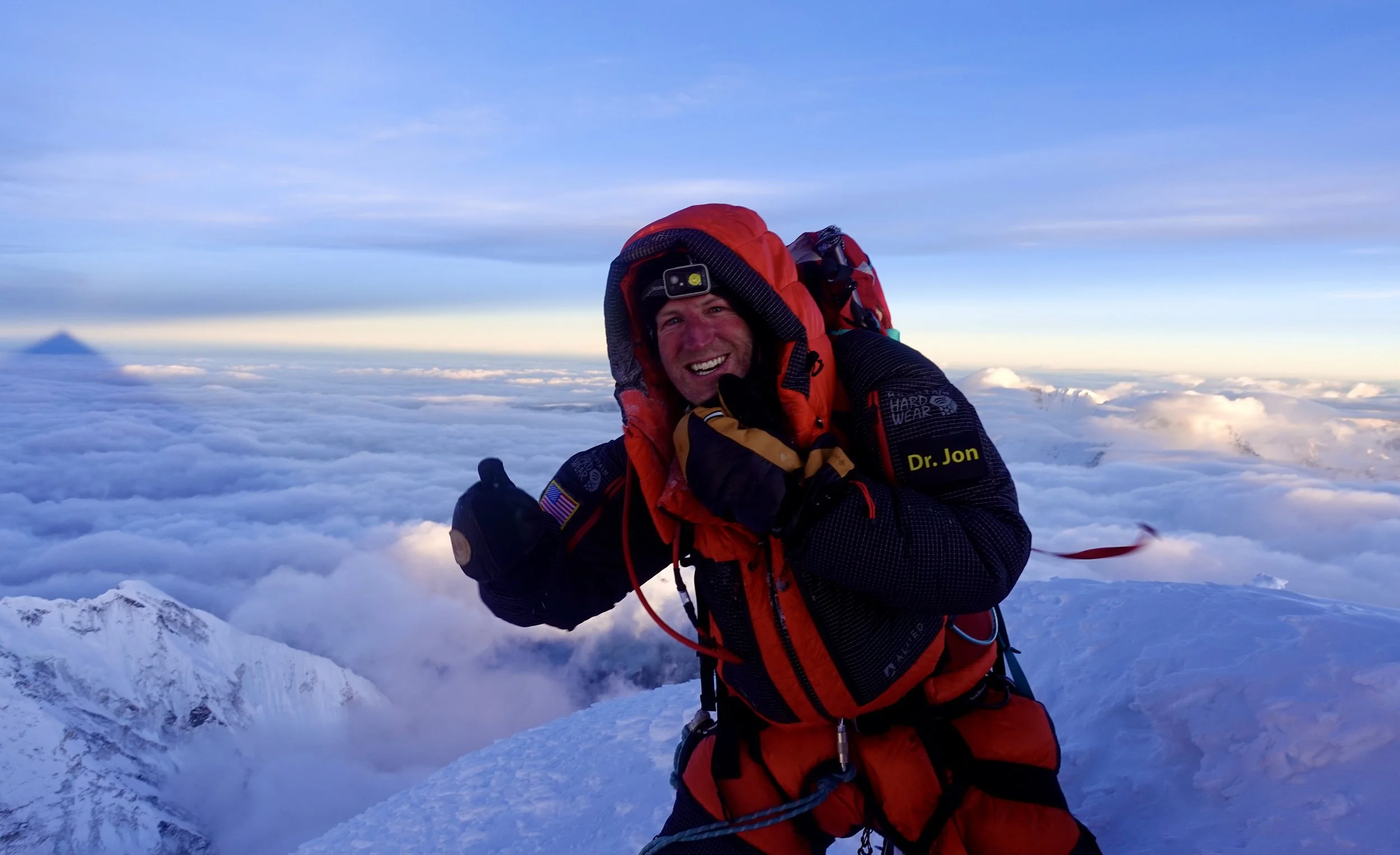 A man dressed in mountaineering gear, smiling, giving a thumbs-up, standing on a snowy mountain peak above the clouds with a view of a distant mountain in the background.
