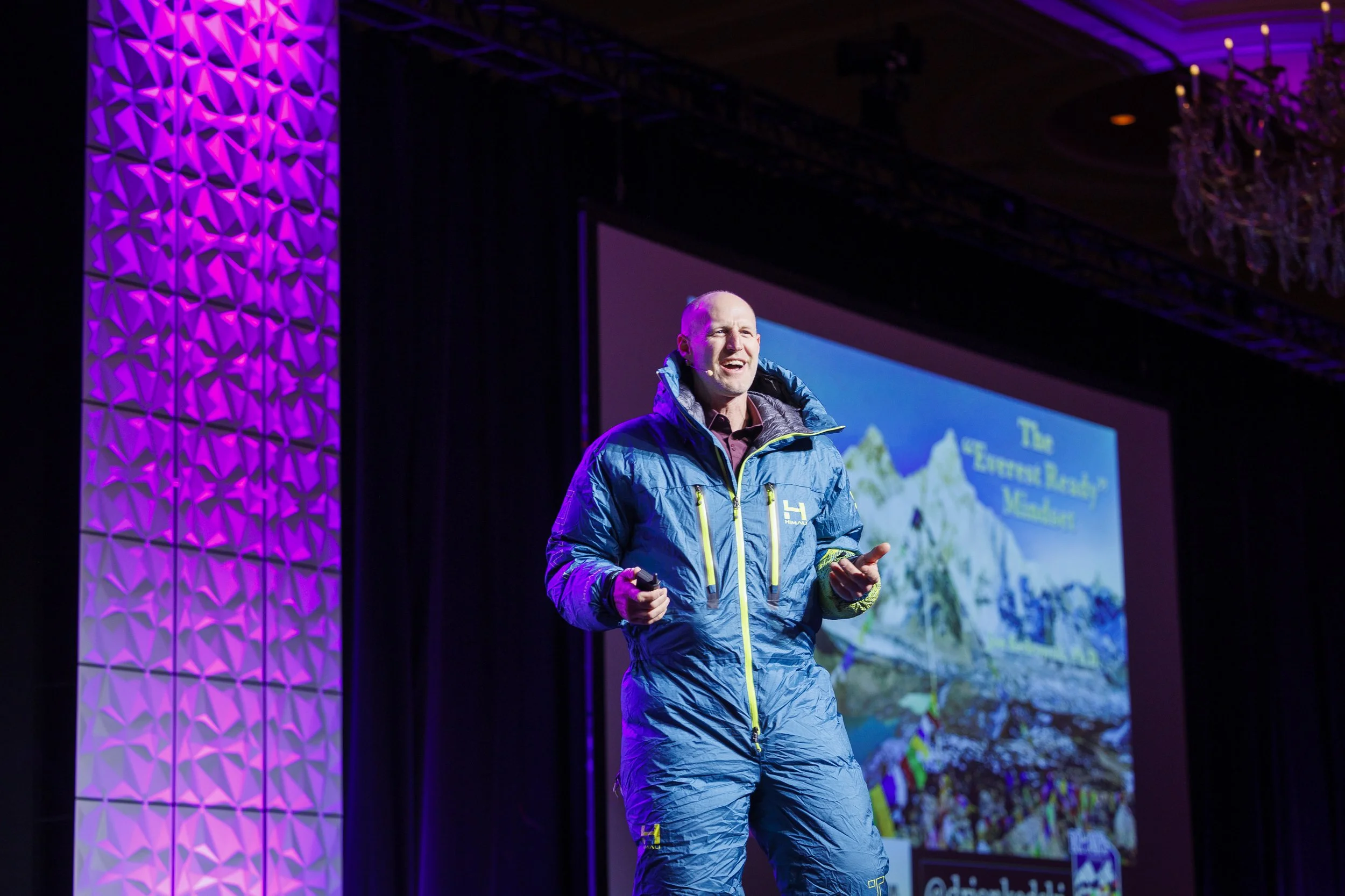 A man in a blue jacket giving a presentation on a stage with a colorful purple-lit background and a large screen displaying mountains and text.
