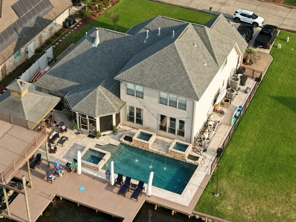 Aerial view of a backyard with a swimming pool, hot tubs, and a wooden deck, adjacent to a two-story house with a gray roof and white exterior, in a residential neighborhood.