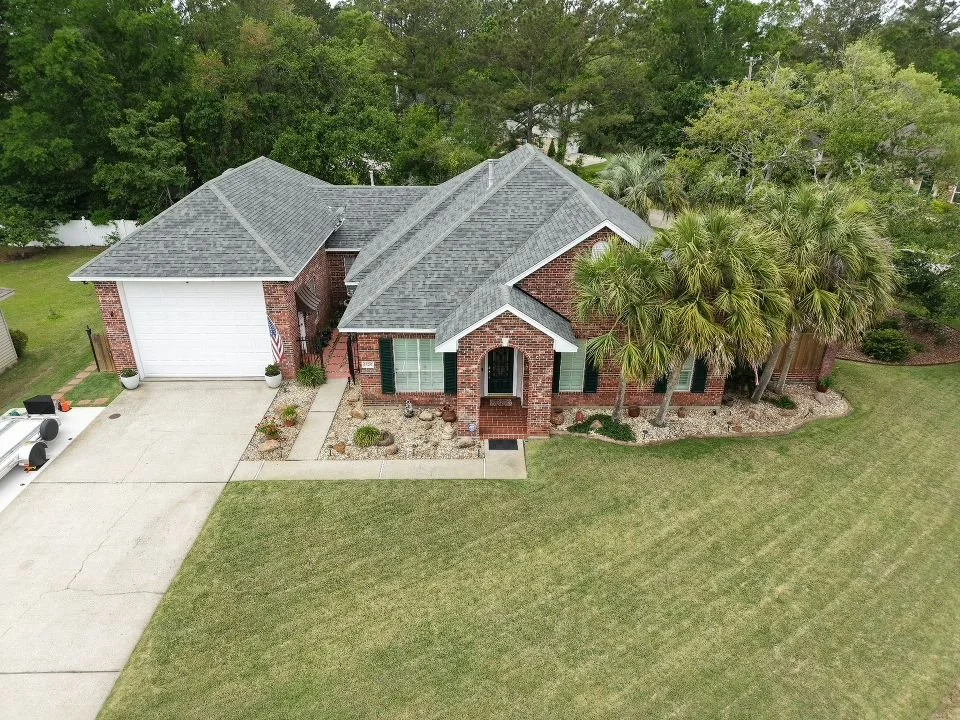 Aerial view of a brick house with a gray shingled roof, green shutters, and a front porch, surrounded by a lawn, trees, and shrubbery.