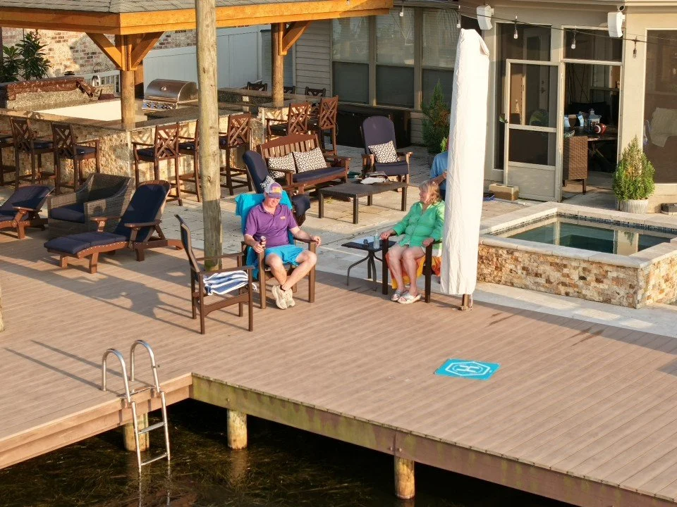 A backyard patio overlooking a body of water with outdoor furniture, including chairs, a table, and a built-in hot tub. Two people are sitting and talking under a covered area with string lights overhead.