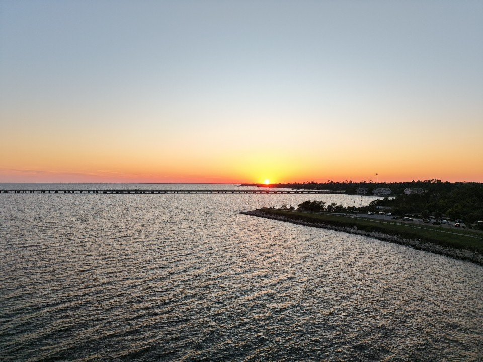 Sunset over a body of water with a bridge in the distance and a shoreline with trees and cars.