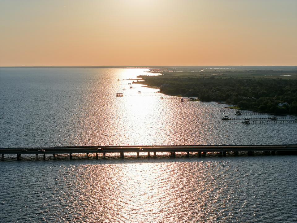 Aerial view of a river with a bridge crossing over it during sunset, with boats on the water and a tree-lined shoreline.