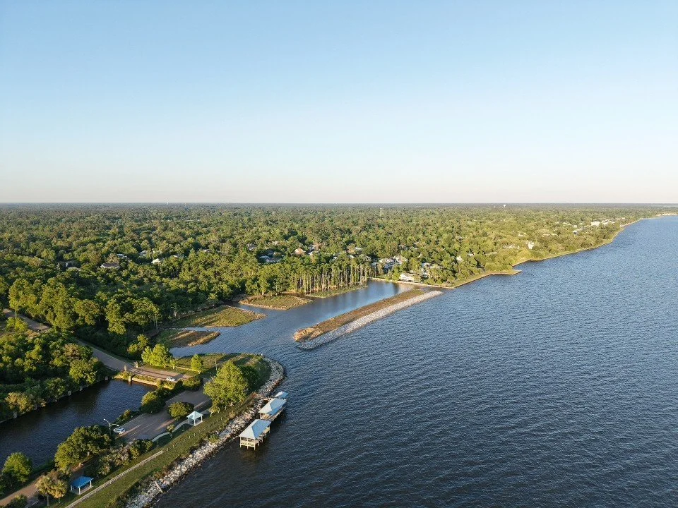 Aerial view of a shoreline with trees, houses, and boat docks along a large body of water under a clear blue sky.