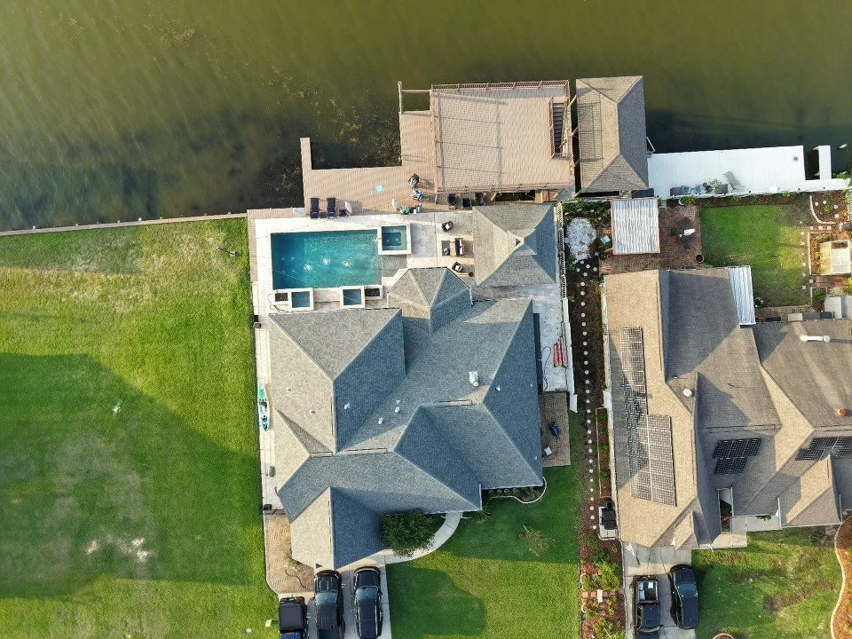 Aerial view of a house with a gray roof, swimming pool, and backyard, situated near a water body with a dock, neighboring houses, and parked cars.