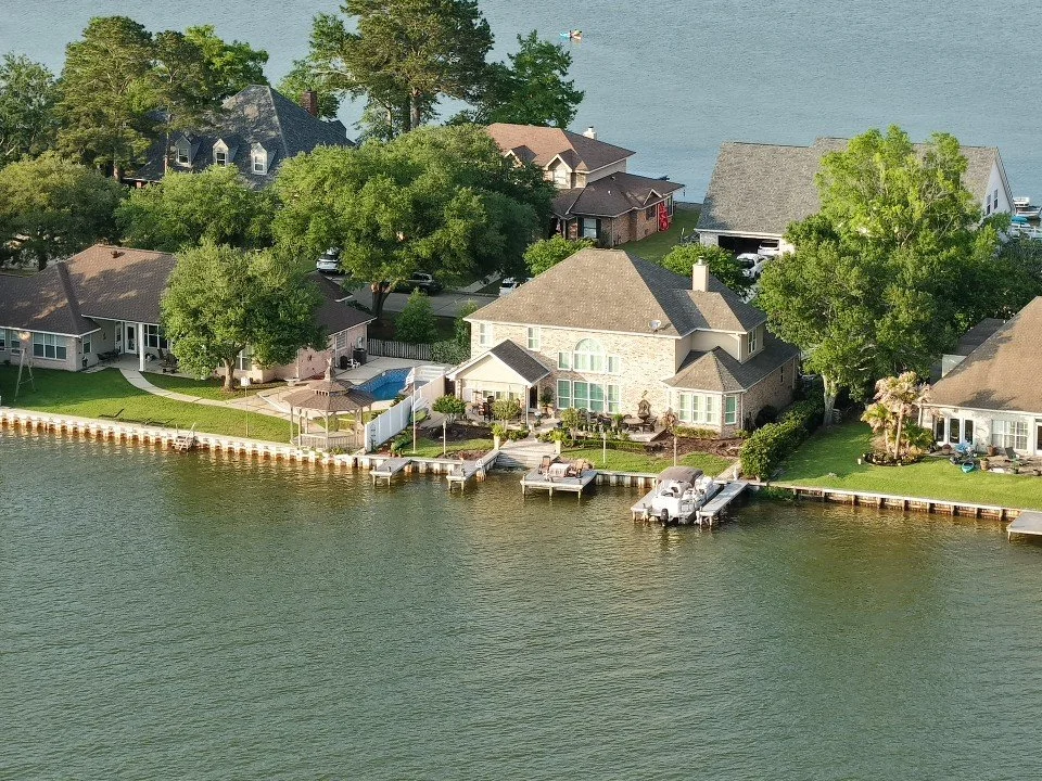 Aerial view of lakeside homes with docks, trees, and a watercraft on a calm body of water.