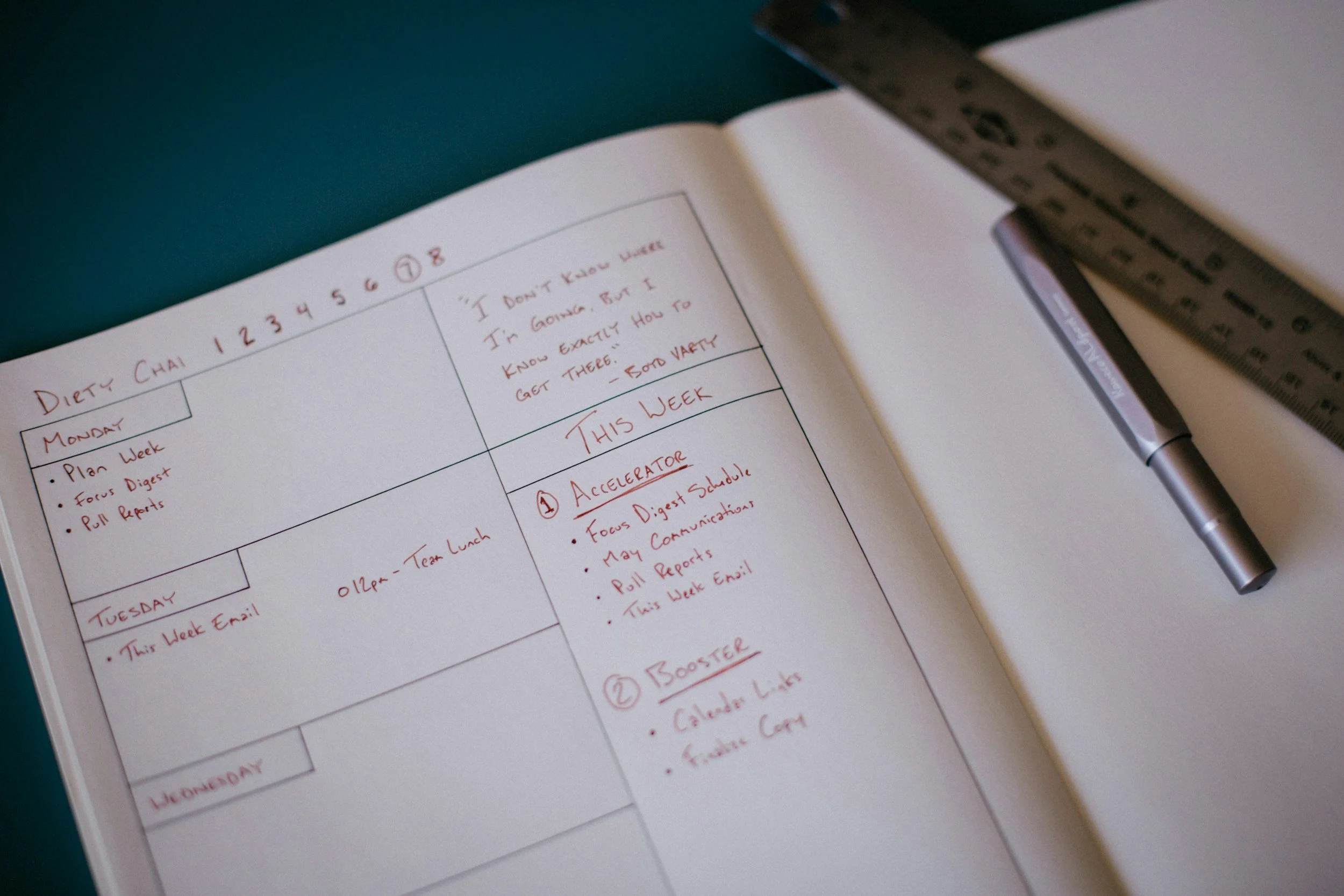A planner or notebook with a weekly schedule written in red ink, a black pen placed on the open page, and a ruler crossing the top right corner of the page. The schedule includes days of the week and notes for each day.