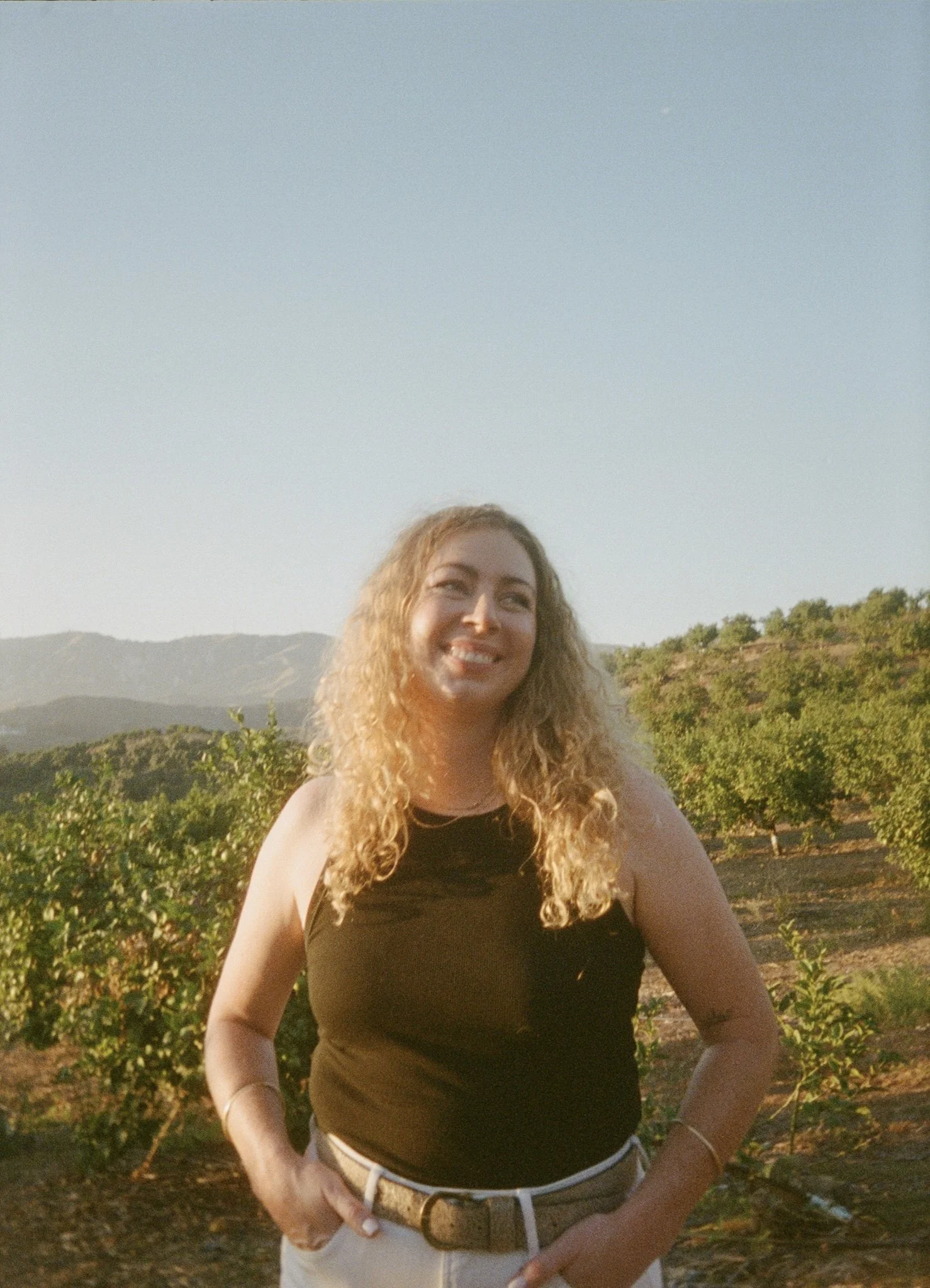 A woman with curly strawberry blonde hair smiling outdoors in an orchard during sunset, wearing a black tank top and cream pants with a belt.