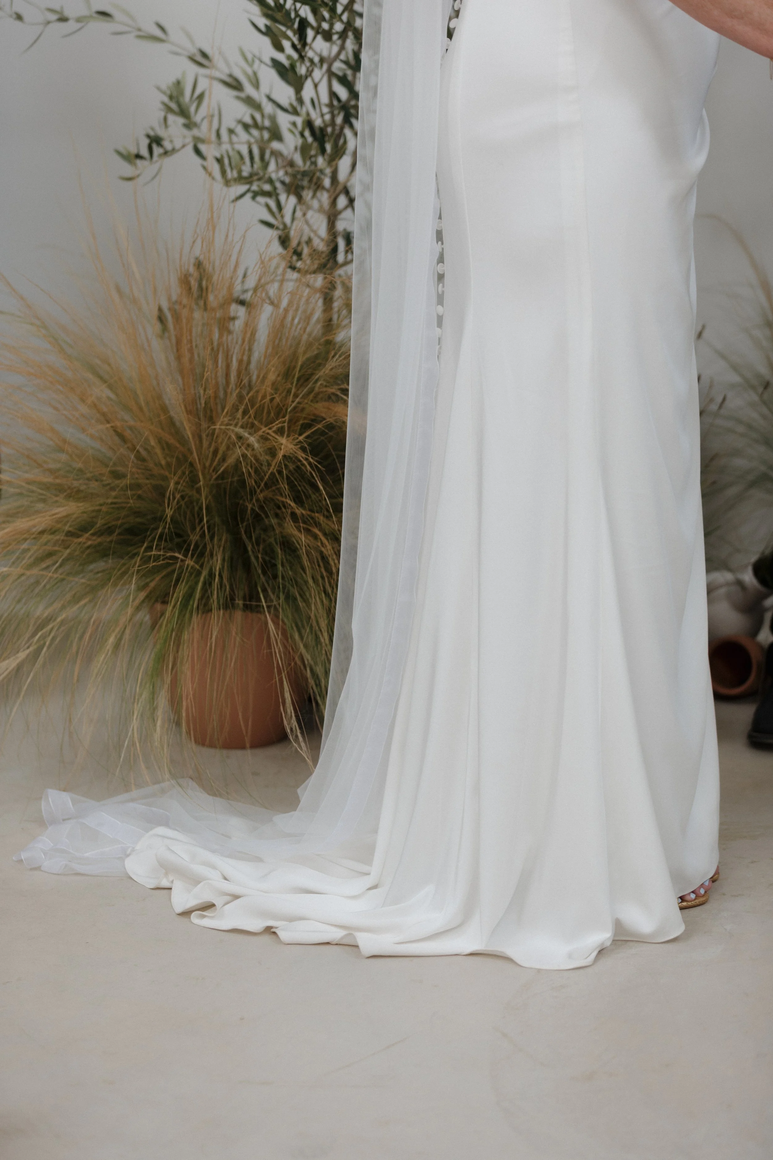 Close-up of a person dressed in a white gown with a flowing train, standing on a light-colored floor next to a large potted plant with long, wispy leaves.
