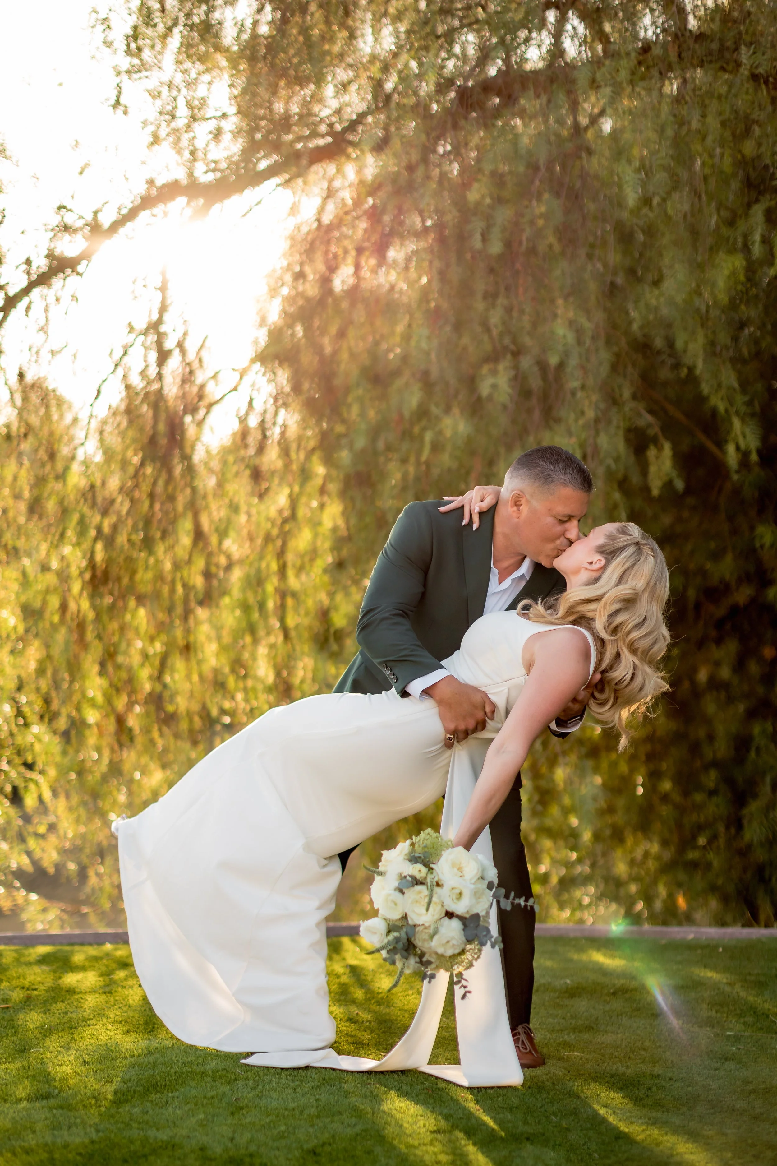 A groom dips and kisses the bride outdoors during sunset, with trees and green grass in the background. The bride holds a bouquet of white flowers.