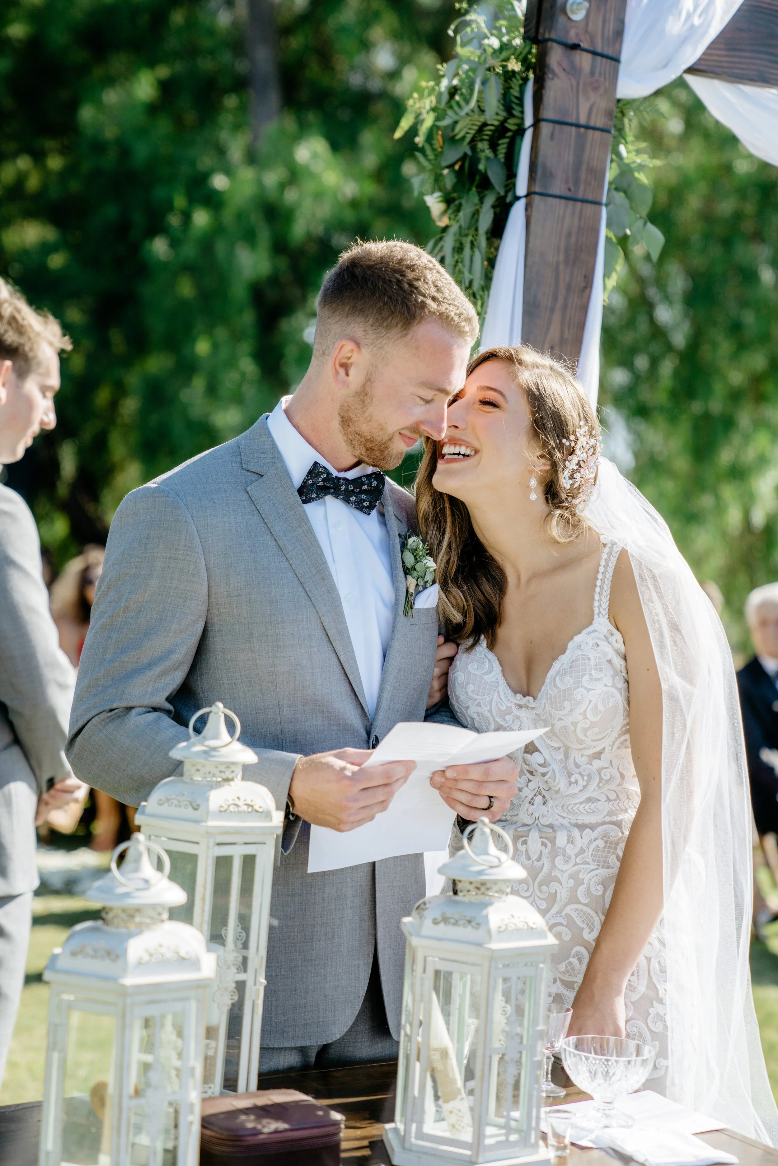 A bride and groom sharing a moment during their outdoor wedding ceremony, with the groom reading a handwritten speech from a piece of paper and the bride smiling.