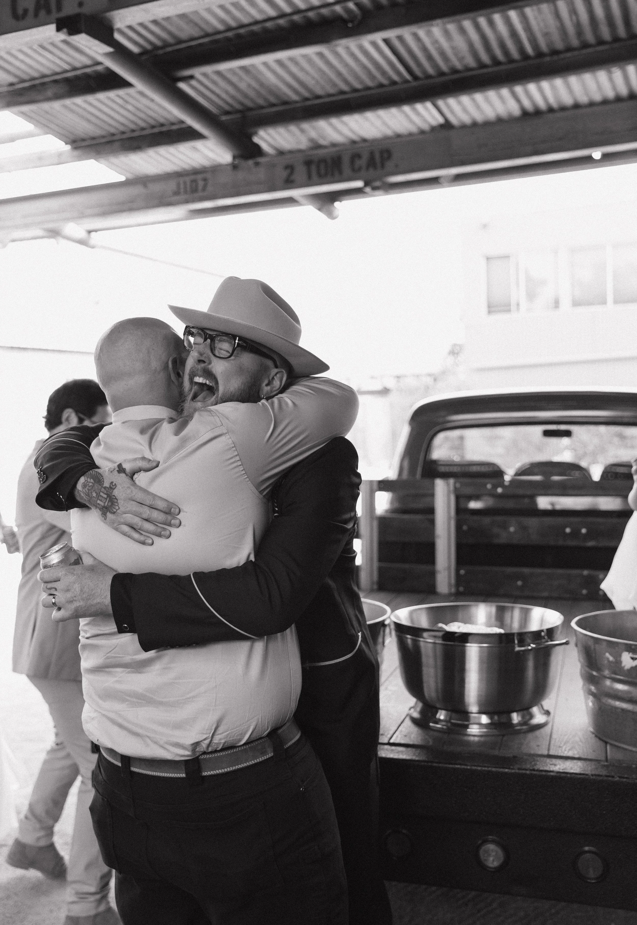Two men hugging at a barbecue restaurant or outdoor event, with a man wearing glasses and a wide-brimmed hat visible. In the background, metal tables, containers, and a truck are visible. The photo is in black and white.