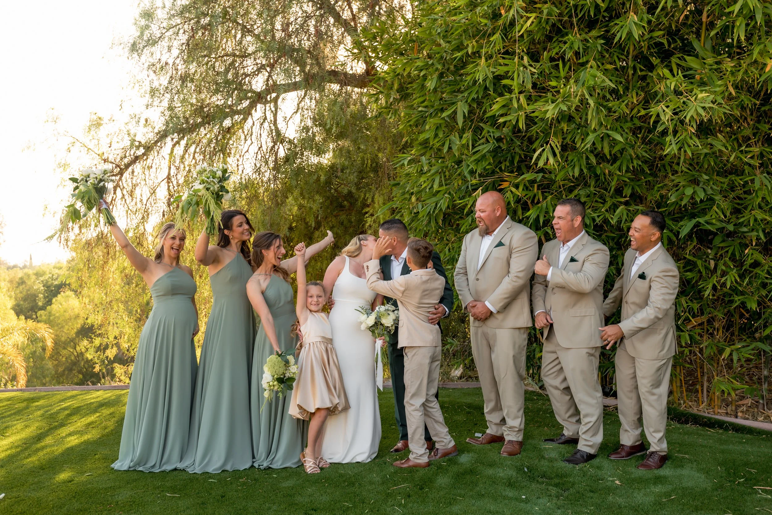 A wedding celebration outdoors with the bride and groom kissing, surrounded by friends and family. The bridesmaids wear green dresses, and the groomsmen wear beige suits. The group is smiling and laughing, holding bouquets, with a lush green background and sunlight filtering through trees.