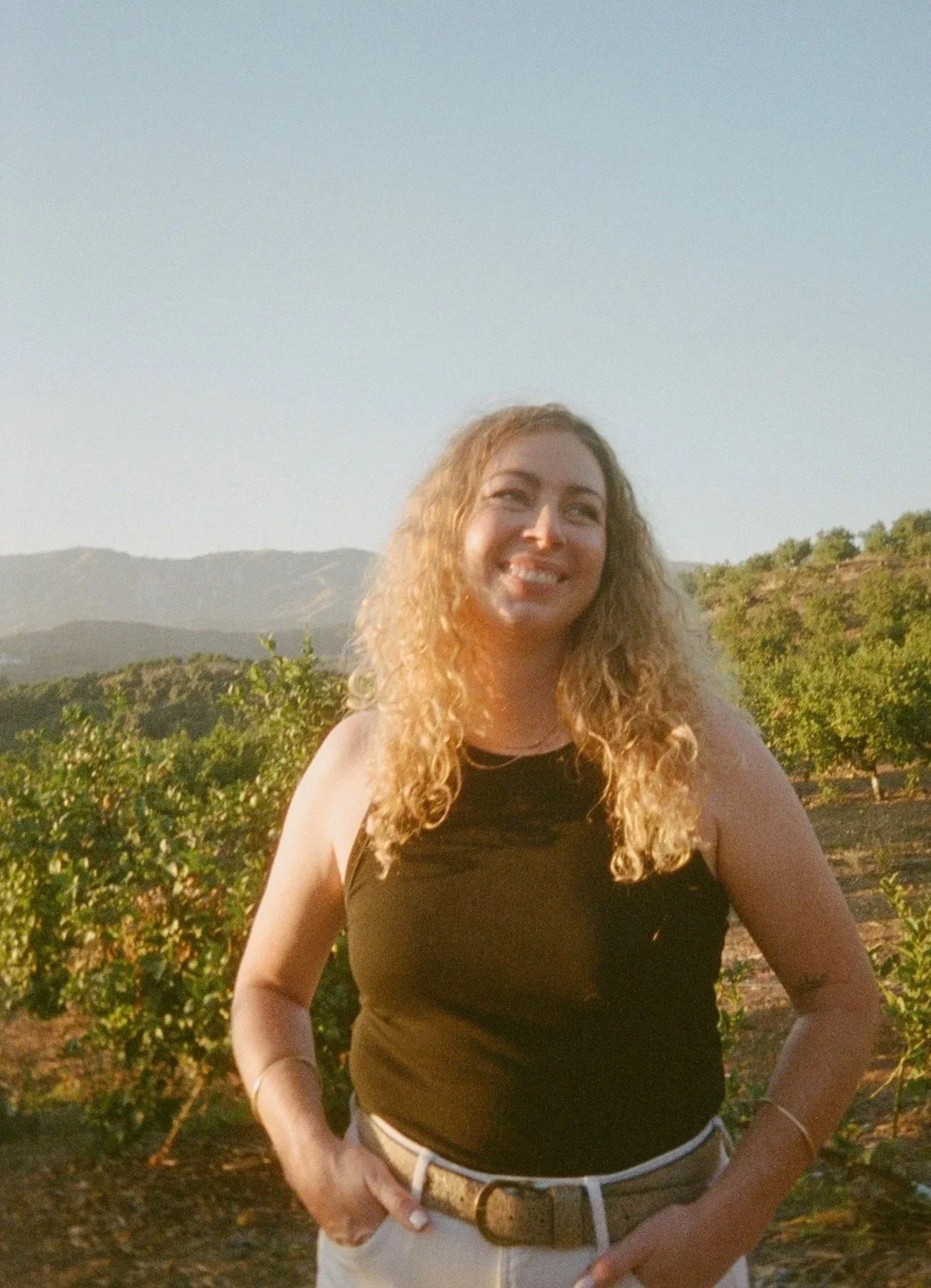 A woman with curly blonde hair standing outdoors with mountains and greenery in the background during sunset, smiling and wearing a black sleeveless top.
