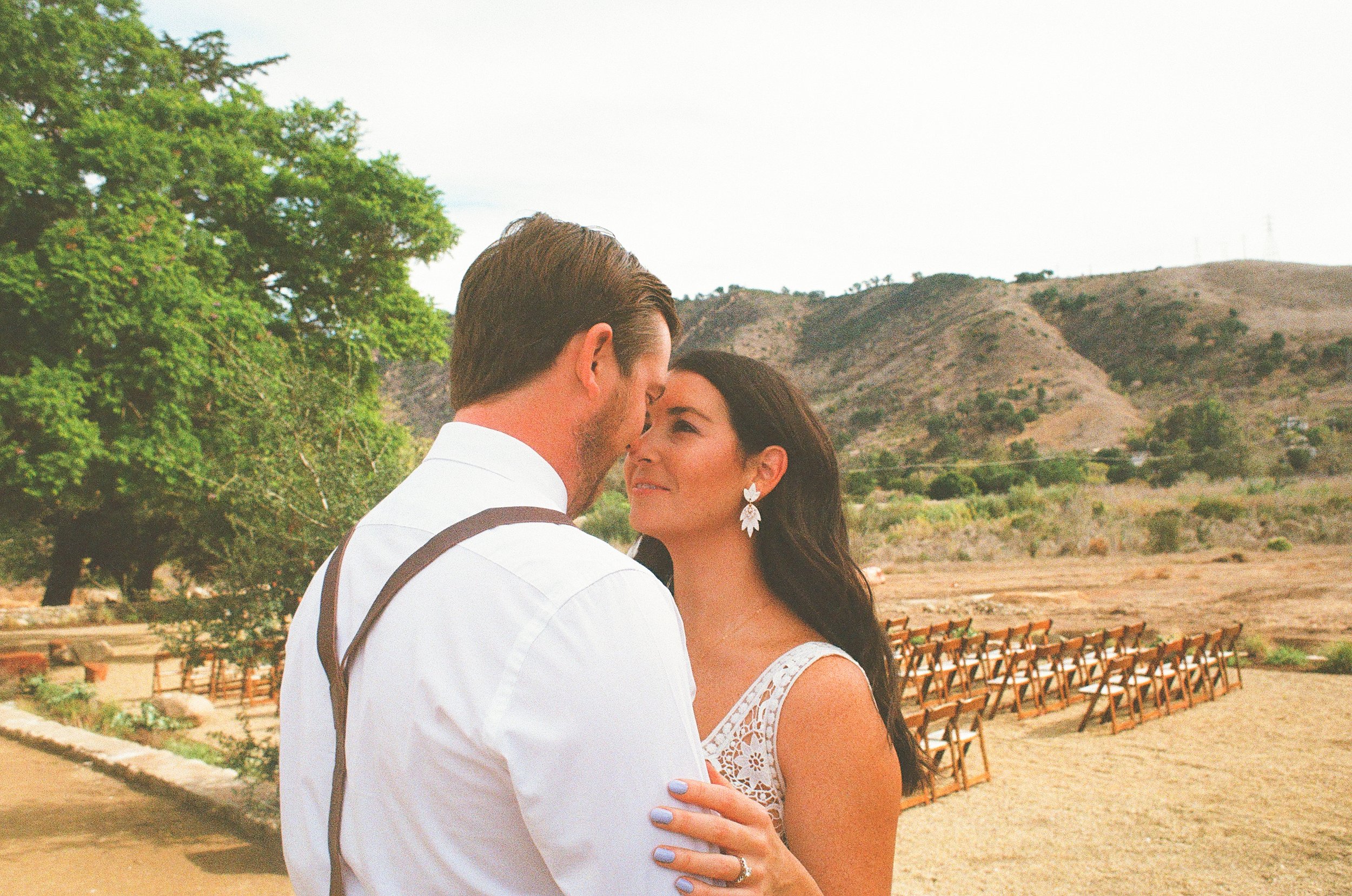 A couple dressed in wedding attire embracing outdoors near a scenic hillside and rows of chairs, with trees and a partly cloudy sky in the background.
