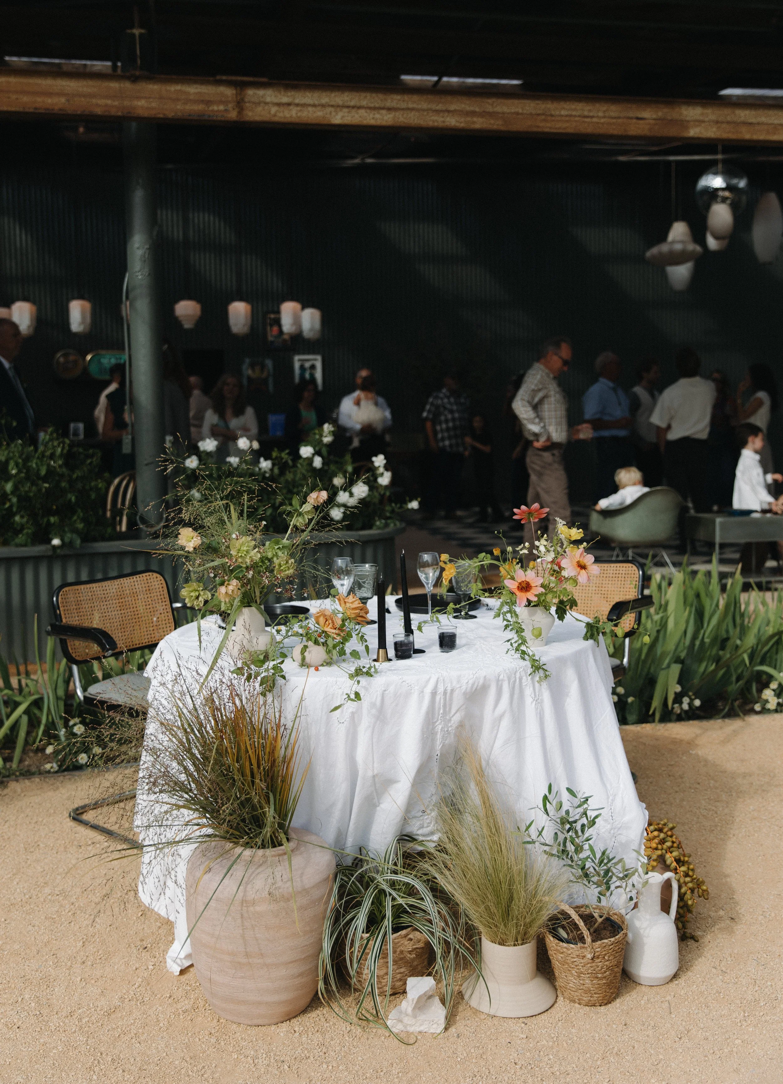 A round table with a white tablecloth, decorated with flower arrangements and surrounded by chairs, in an indoor or semi-outdoor setting, with potted plants in the foreground and a group of people in the background.