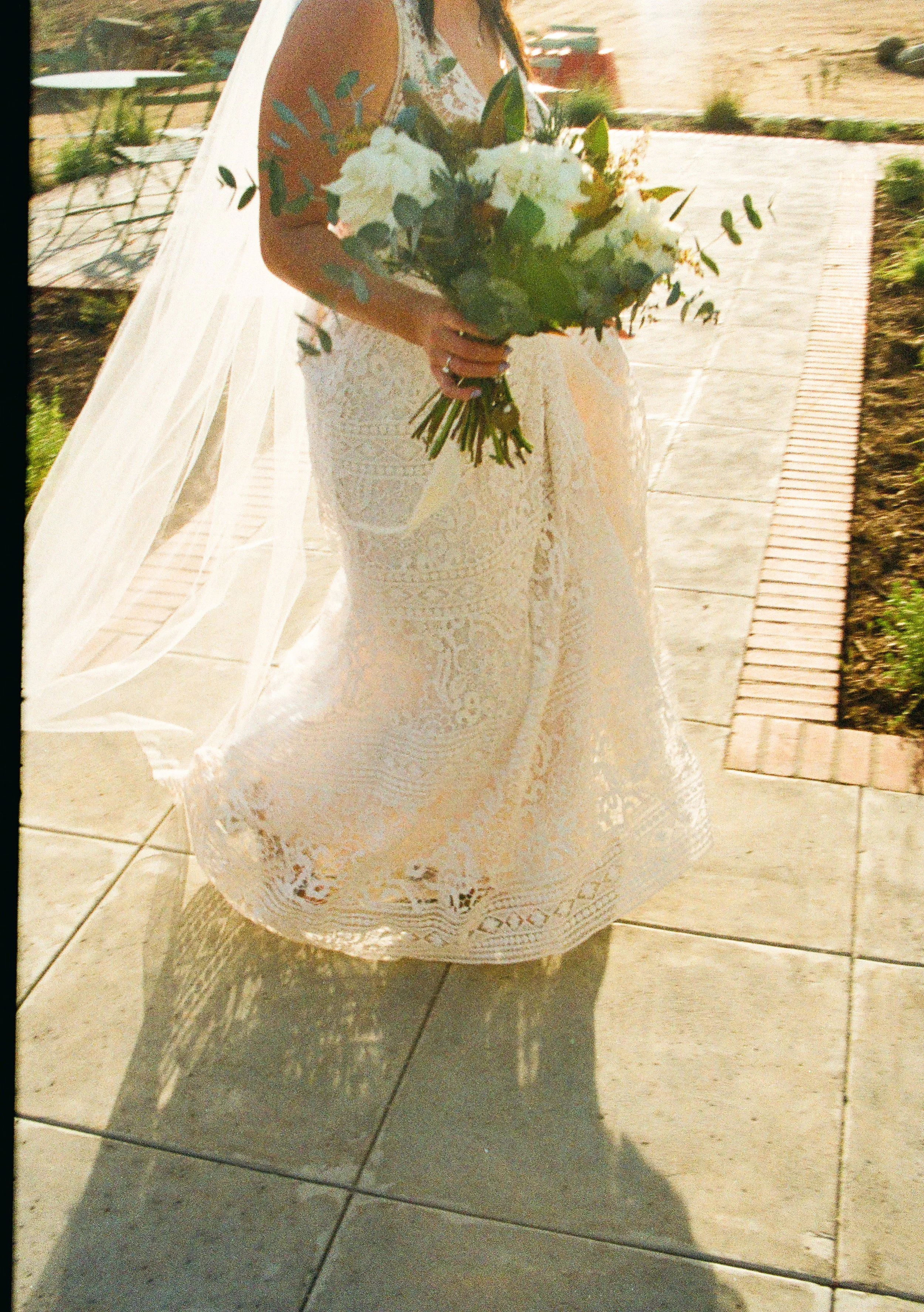 A woman in a white lace dress holding a bouquet of white flowers and greenery, standing outdoors on a paved walkway.