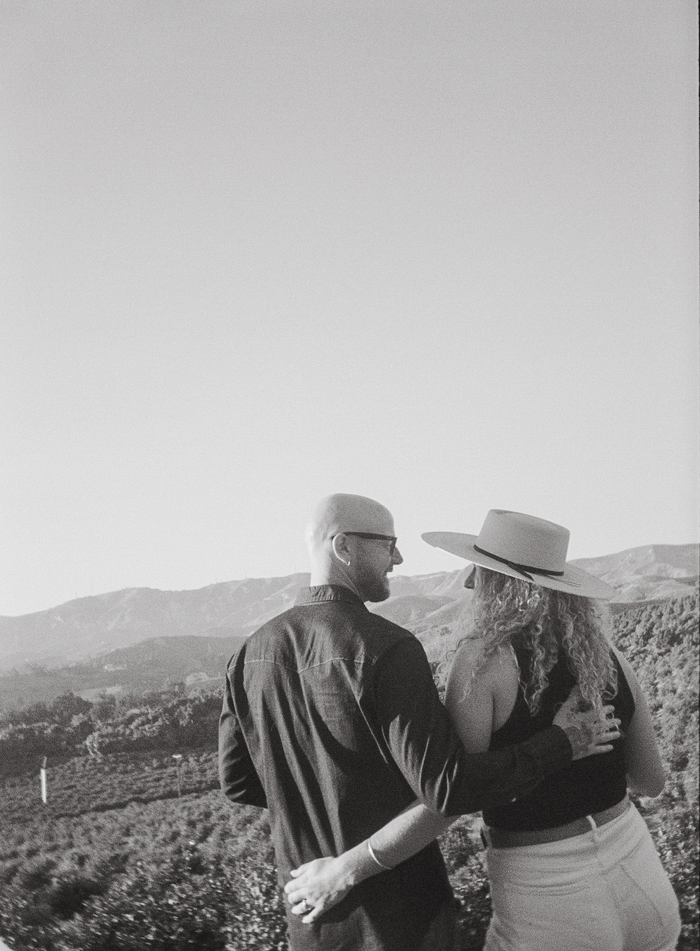 A black-and-white photo of a man and woman standing outdoors in a hilly landscape. The man is wearing glasses and a dark shirt, and the woman is wearing a wide-brimmed hat and light-colored pants. They are facing each other, holding hands, and smiling.