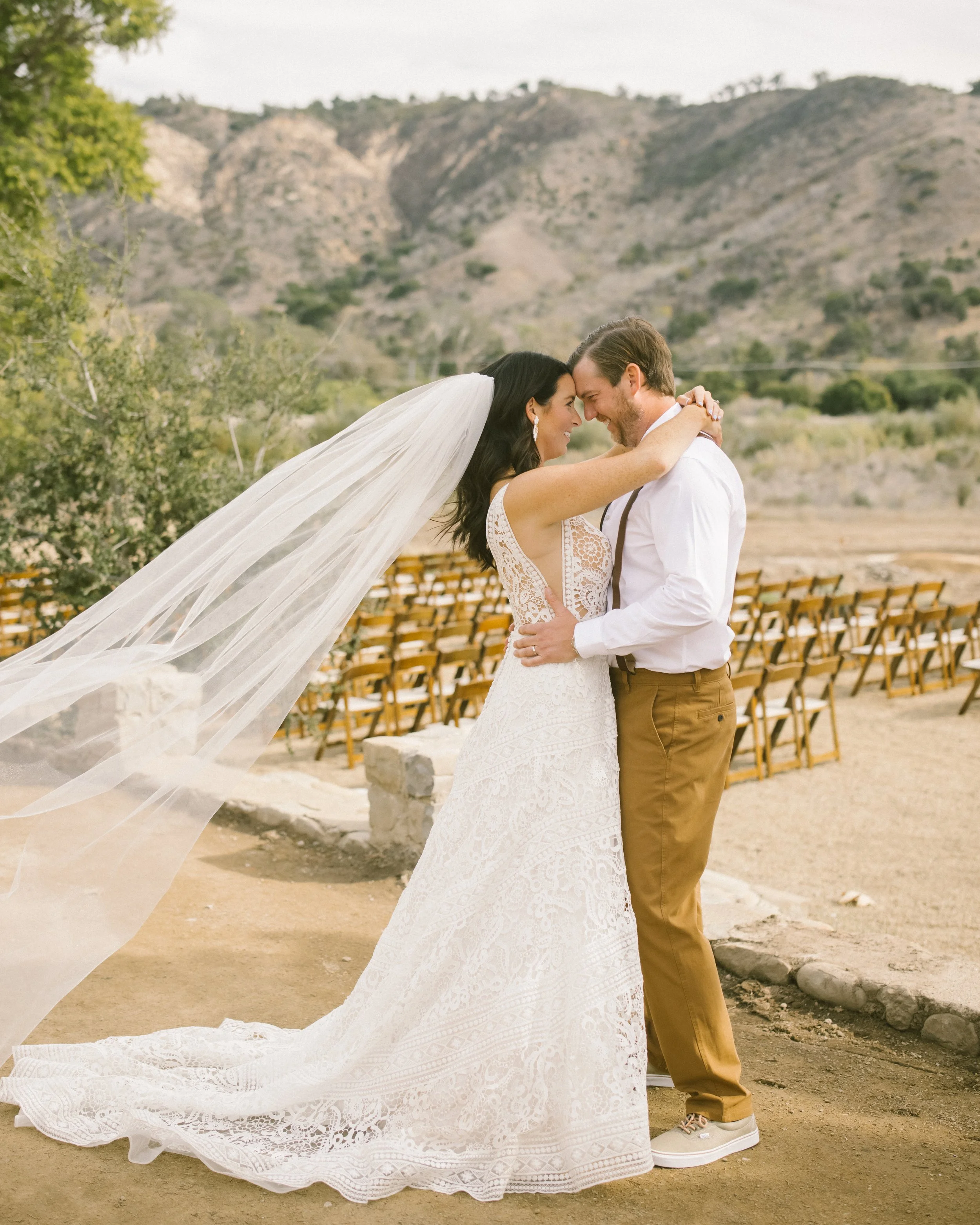 A bride and groom holding each other close, smiling, in an outdoor mountain setting. The bride wears a lace wedding gown and veil, while the groom wears a white shirt and tan pants. Rows of chairs are visible in the background.