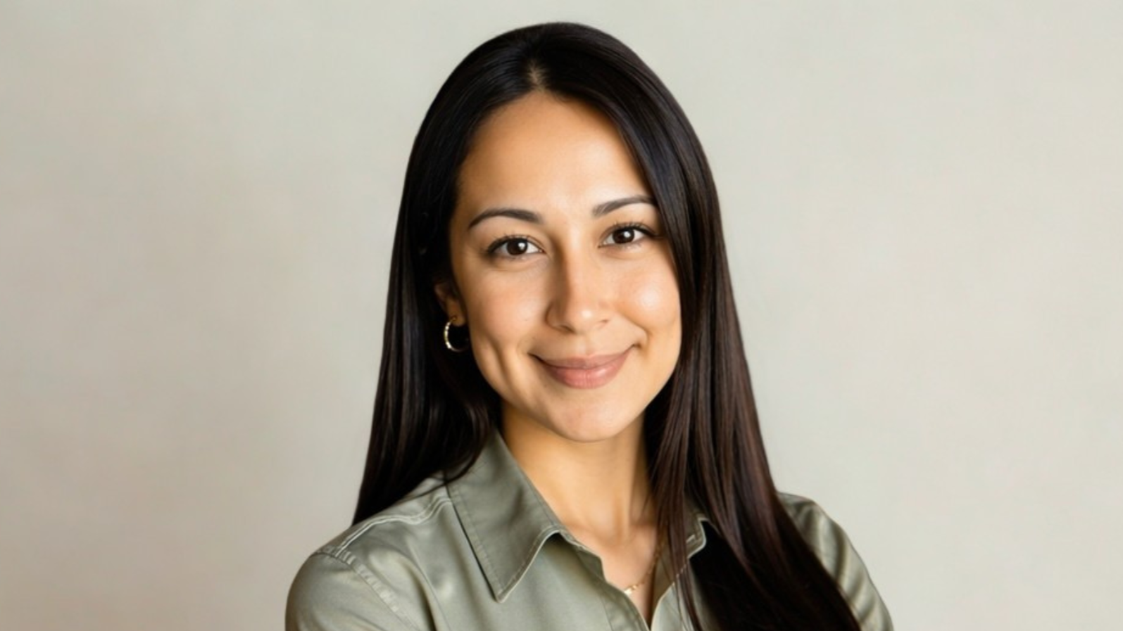 A woman with long dark hair and hoop earrings smiling at camera wearing a light khaki shirt against a plain light background.