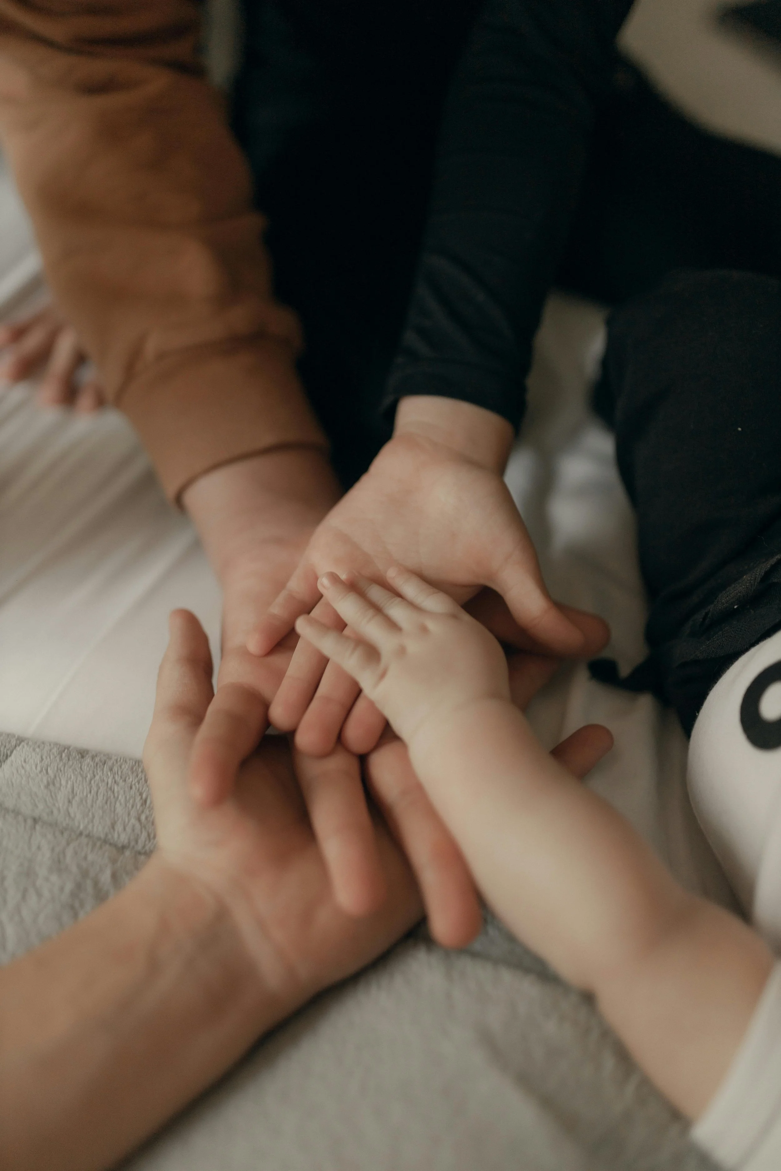 Multiple hands of different sizes, likely an adult and children, are intertwined and resting on a soft surface.