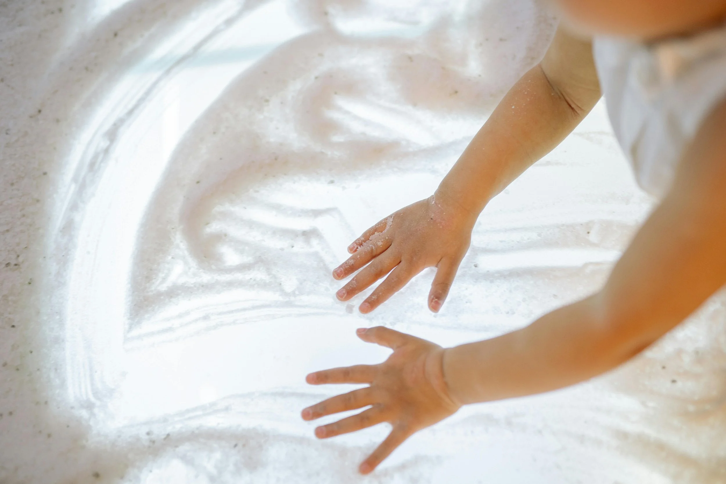 Children's hands are reaching out and touching a white, sandy surface.