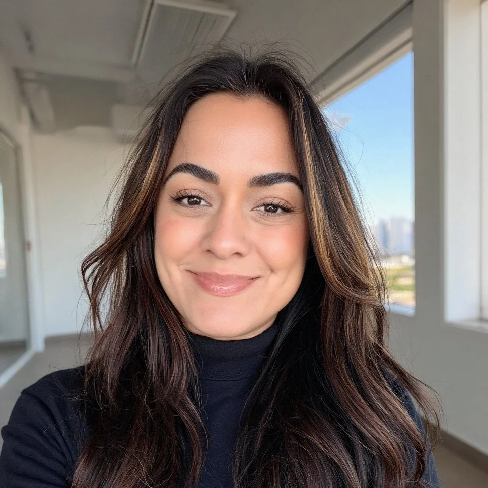 A woman with long, dark hair smiling in a well-lit indoor space with large windows showing a cityscape in the background.