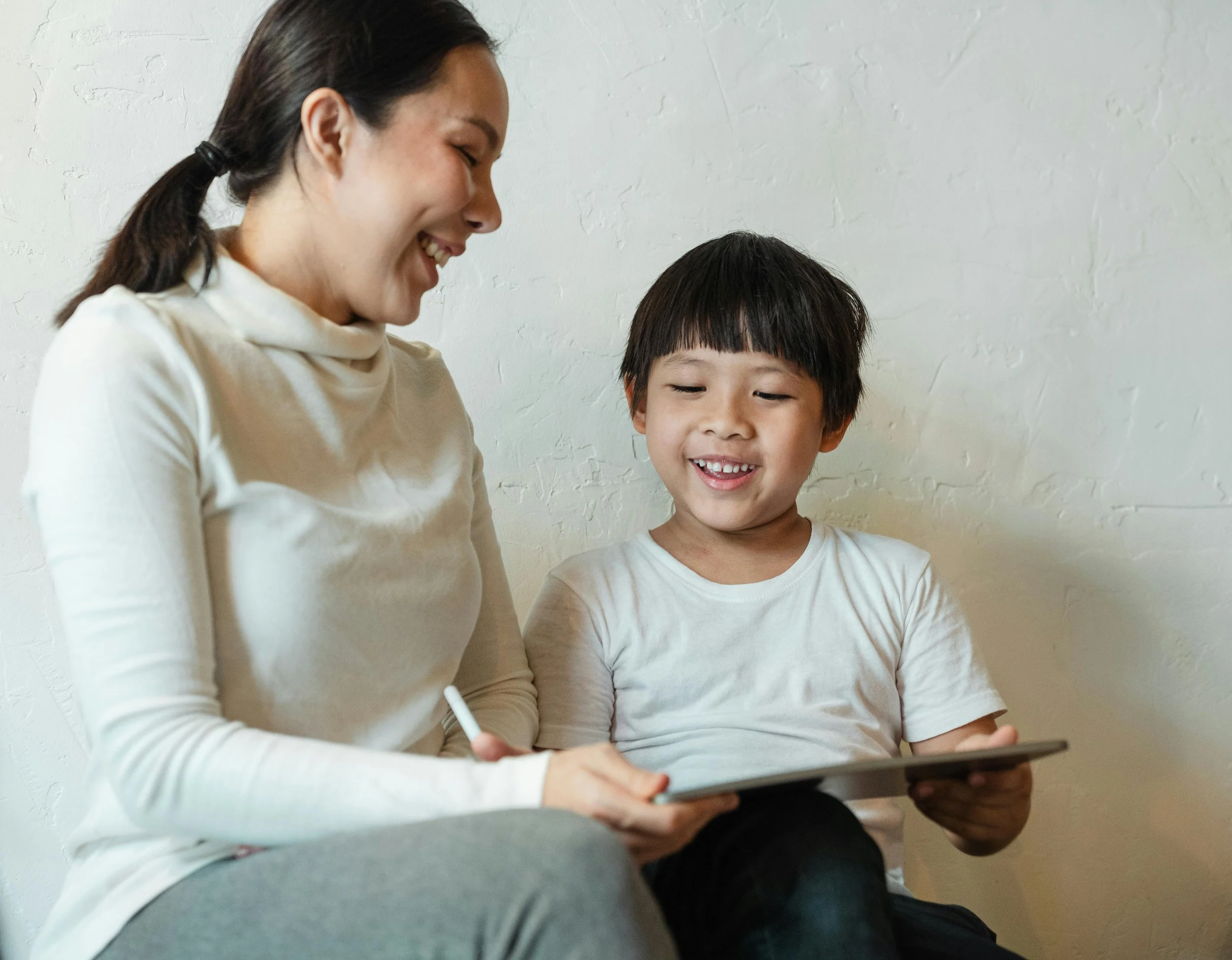 A woman with dark hair tied back smiling while sitting next to a young boy with dark hair, both looking at a tablet device together.