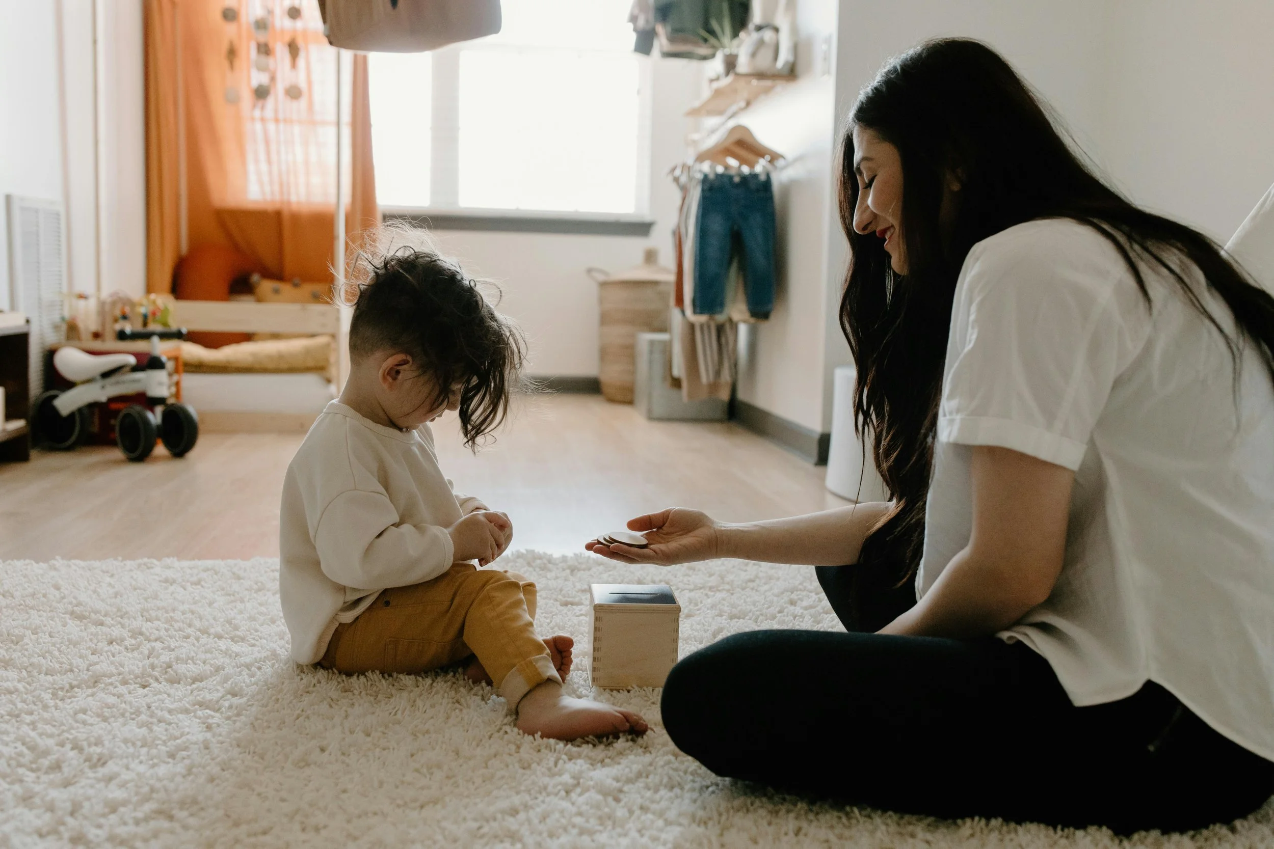 A woman and young girl sitting on a white rug in a bright room, playing a game with wooden blocks. The woman has long dark hair and is smiling, while the girl has curly hair and is focused on the game.