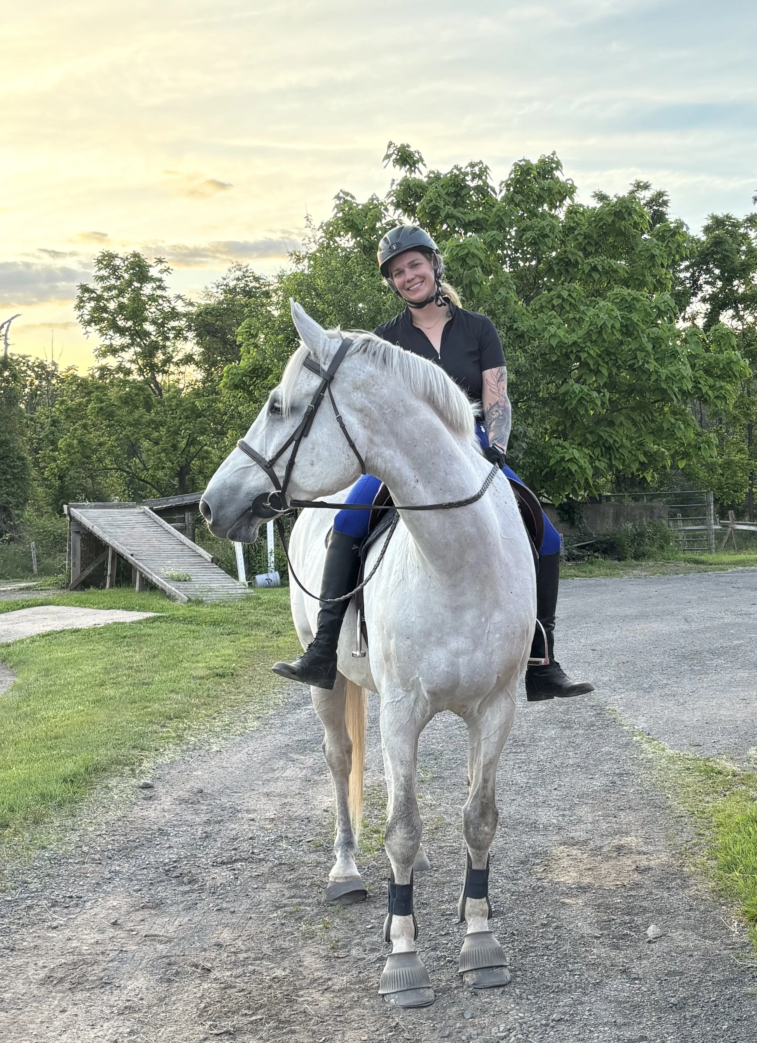 A woman riding a white horse outdoors, wearing a black helmet, black shirt, and black riding boots. The horse has protective boots on its legs. There are trees and a wooden ramp in the background, with a sky filled with clouds at sunset.
