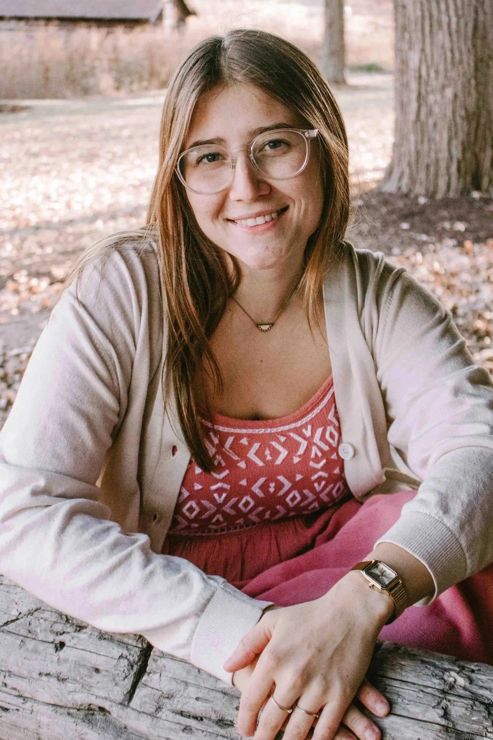 A young woman with glasses and long brown hair sitting outdoors at a wooden table, smiling, with trees and fallen leaves in the background.