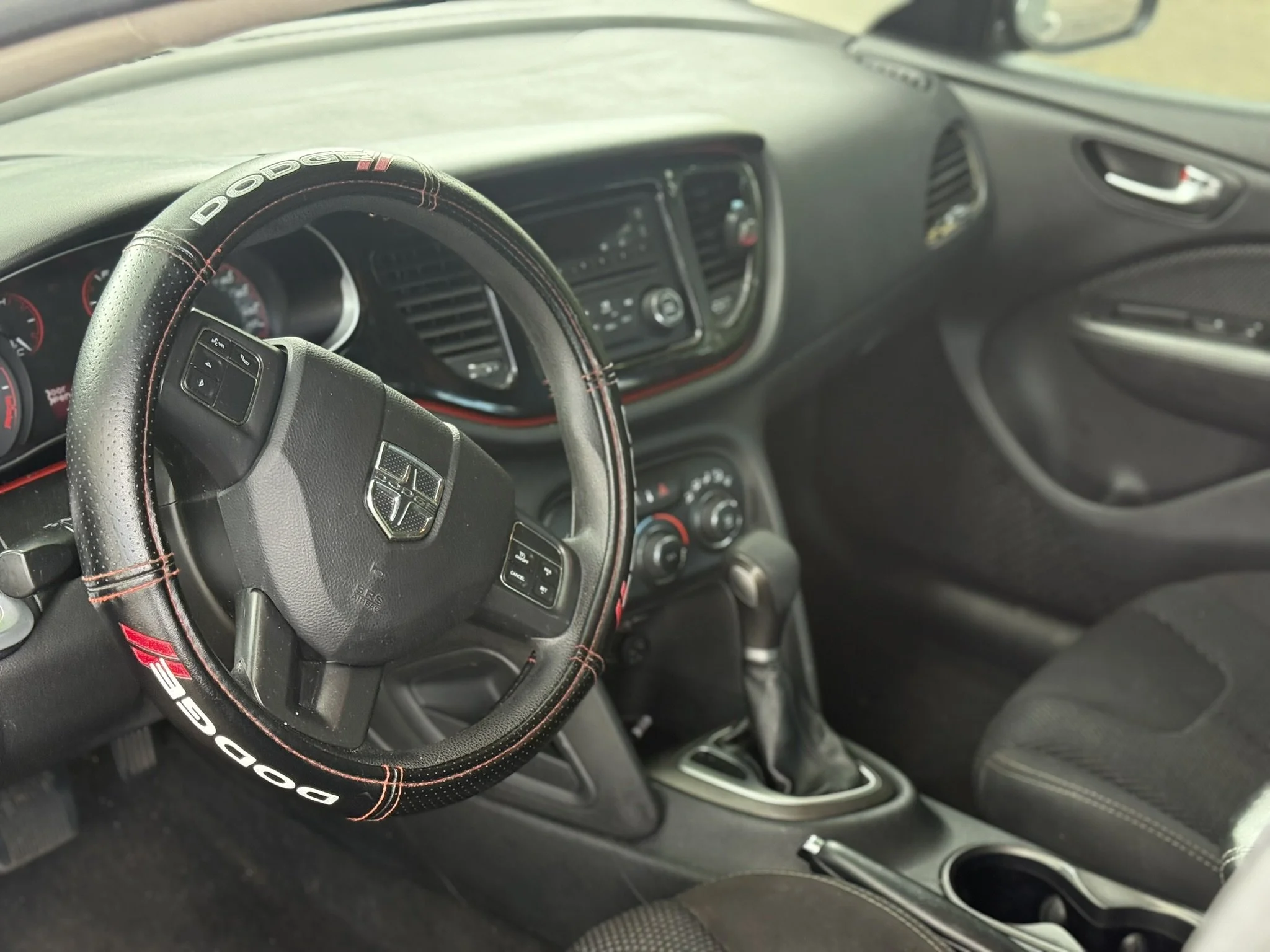 The image shows the interior of a car, focusing on the steering wheel with a Dodge logo, black dashboard with controls and display, and other interior features.