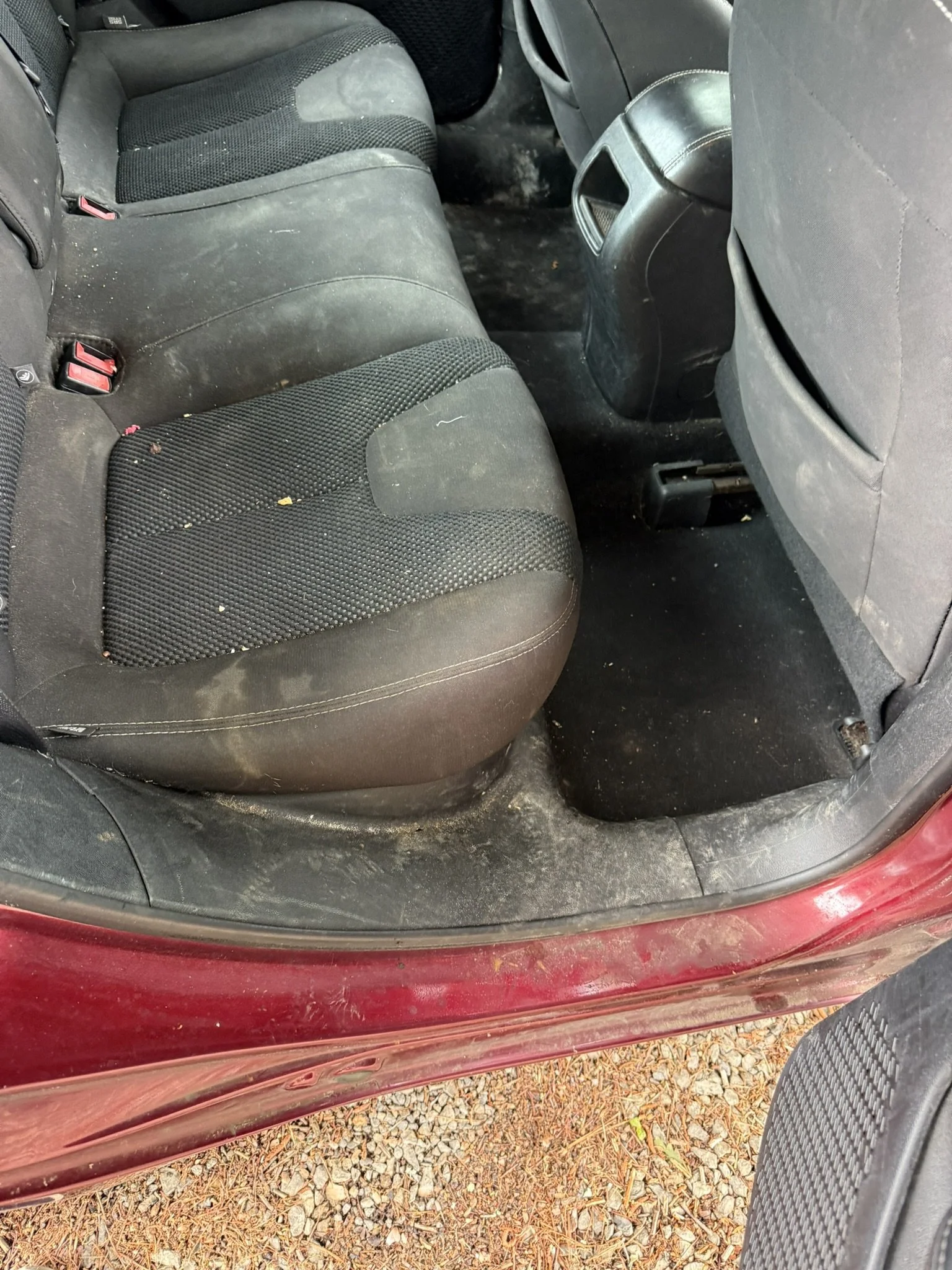 The interior of a red car with a dirty black fabric back seat and floor, showing some trash and a black plastic piece on the floor, viewed from the open door.