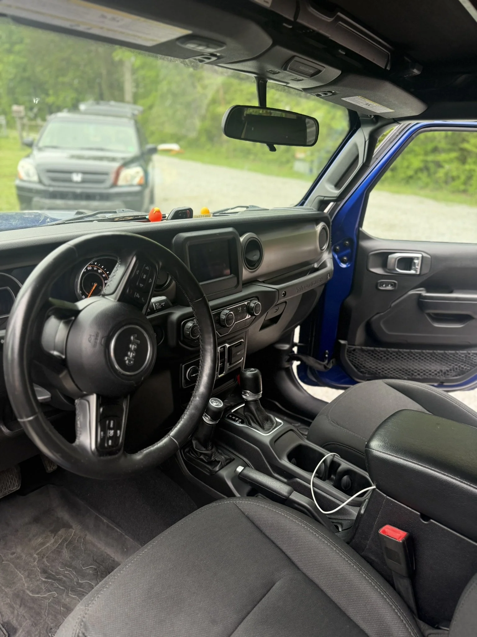 Interior view of a blue off-road vehicle showing steering wheel, dashboard, gear shift, and front passenger seat with grass and trees visible outside through windshield.