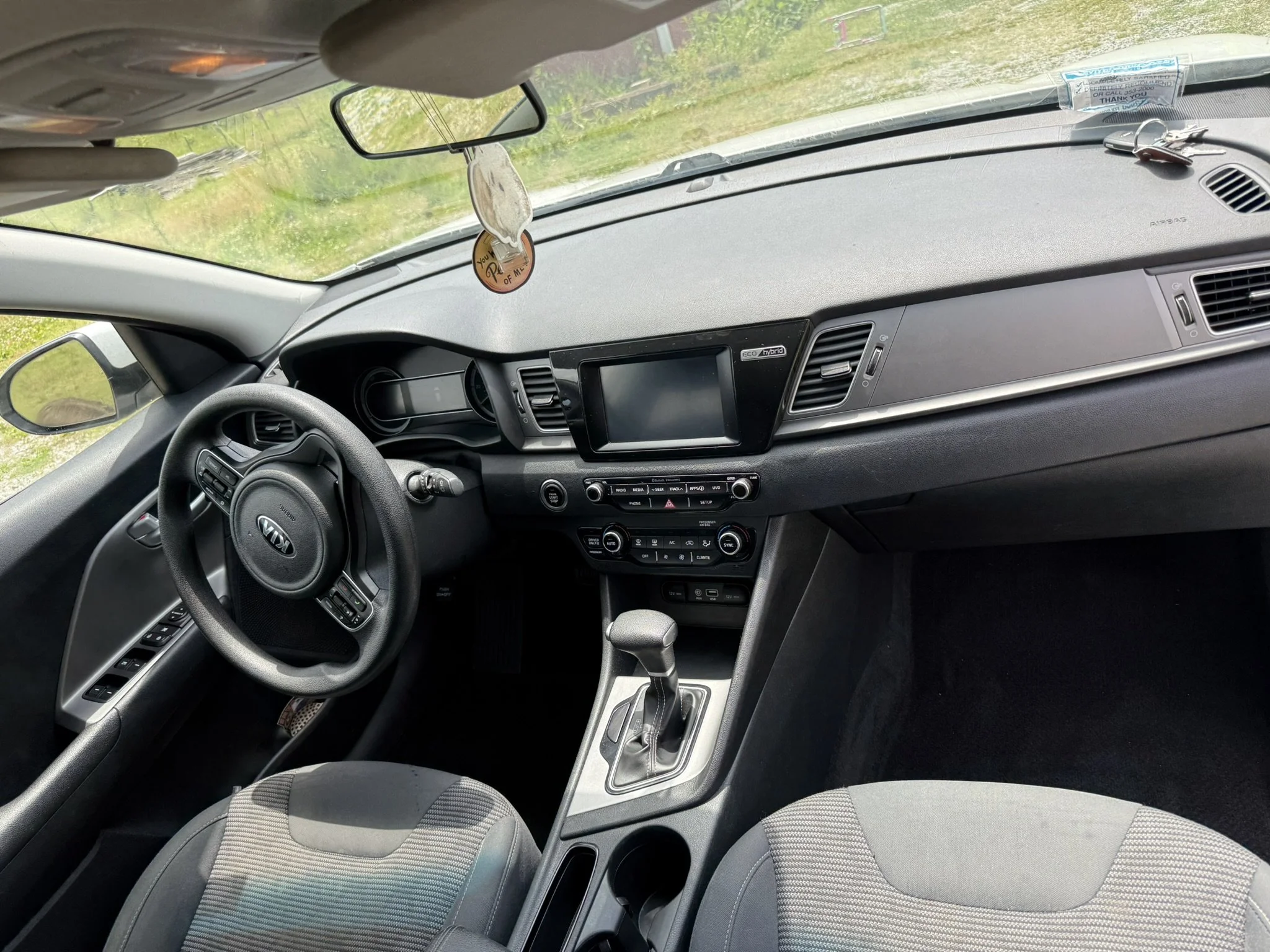 Interior of a car, showing the dashboard, steering wheel, gear shift, and part of the front seats. The dashboard has a digital display and control buttons, with a rearview mirror and dangling decorative items visible.