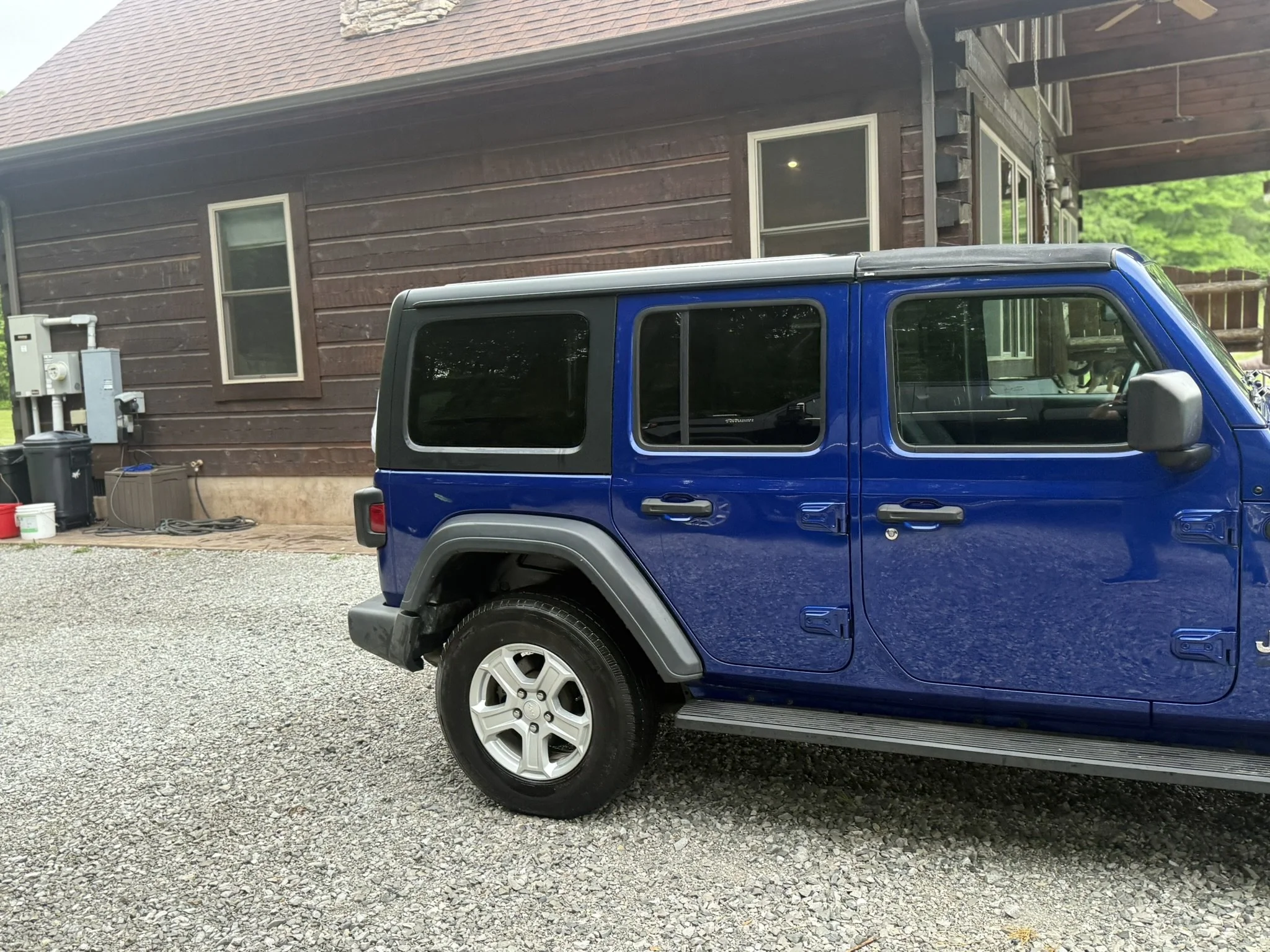 A blue Jeep parked on a gravel driveway in front of a brown wooden house with a sloped roof and multiple windows.